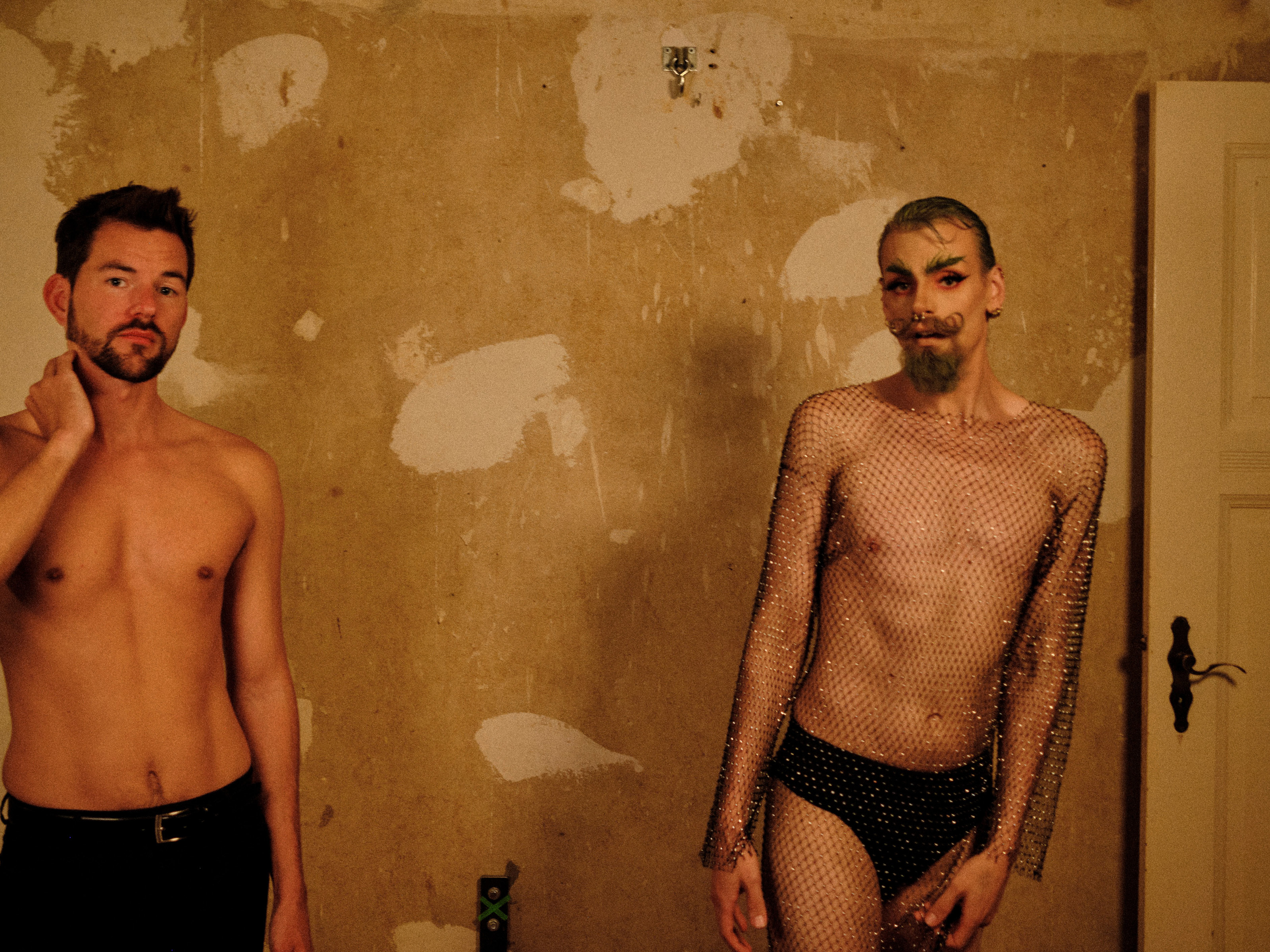 A young man with a bare torso and short brow hair and a short beard with black pants stands in front of an unrenovated wall. Next to him is another young man in black briefs. He is wearing a net dress with fake diamonds. He is wearing intense, dark Make-up and has his beard twirled upwards. His hair is tightly gelled back and his eyebrows are strikingly highlighted.