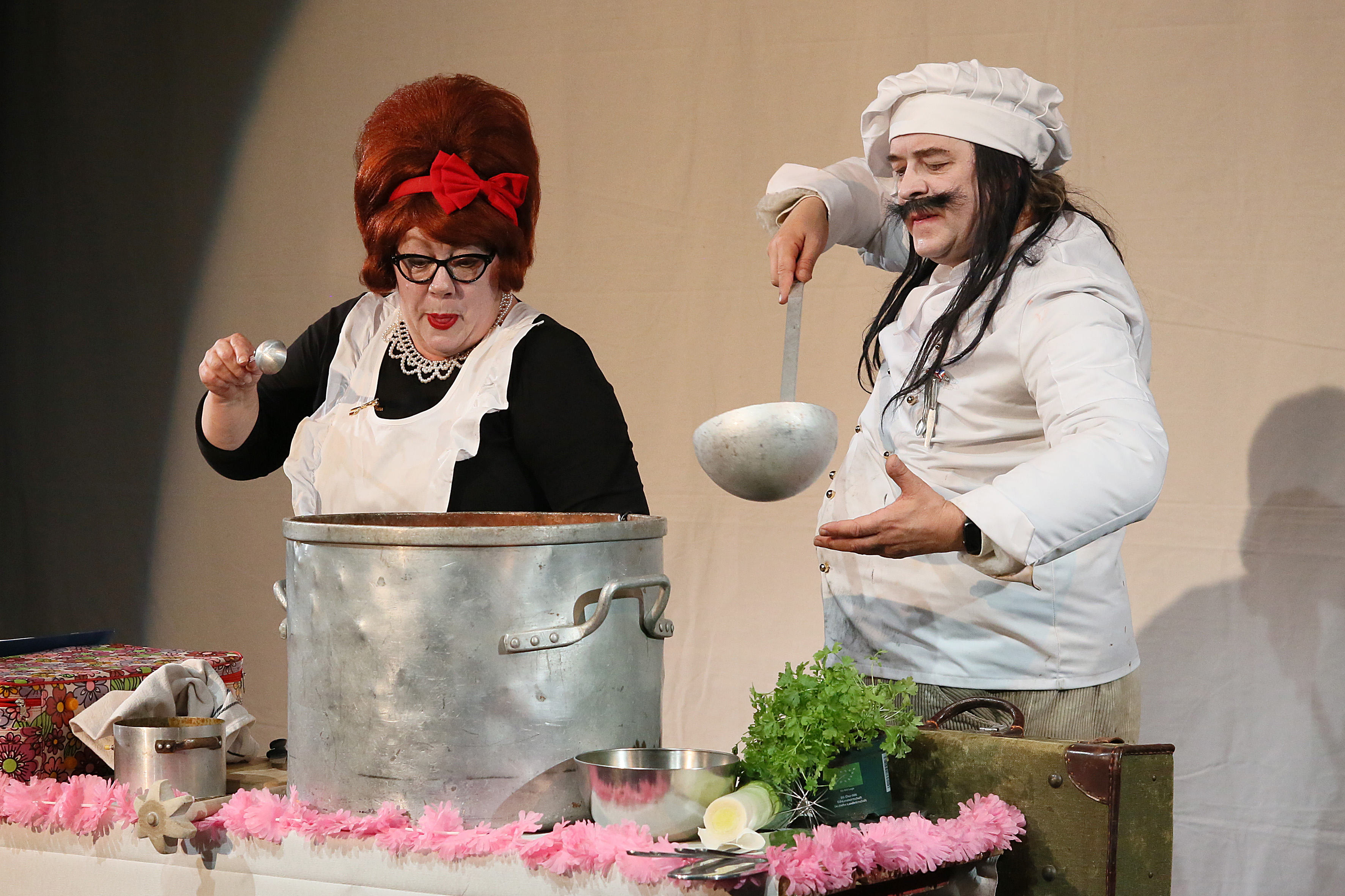 A plump chef with long black hair and a black moustache holds a large silver ladle in his hand. Next to him is a woman with short red hair standing next to a large pot. The pot is placed on a table with fresh vegetables and a pink garland.