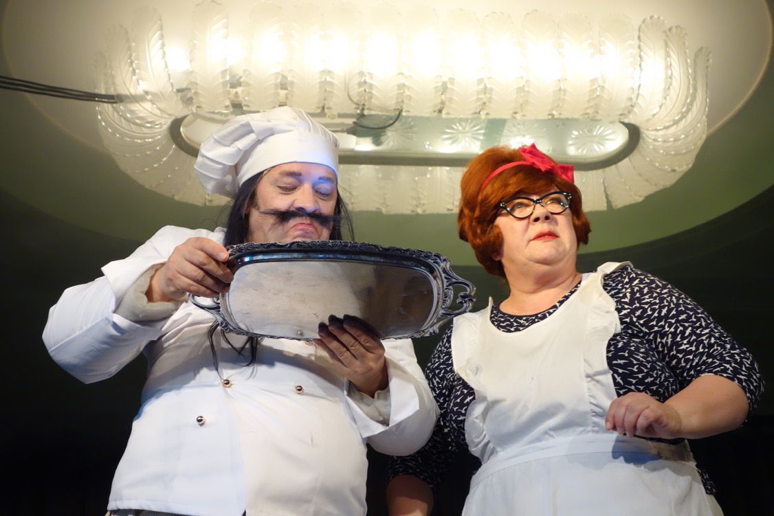 A fat chef with a moustache and chef's hat stands next to a woman with short red hair, holding a silver tray. A large, opulent lamp hangs above them.