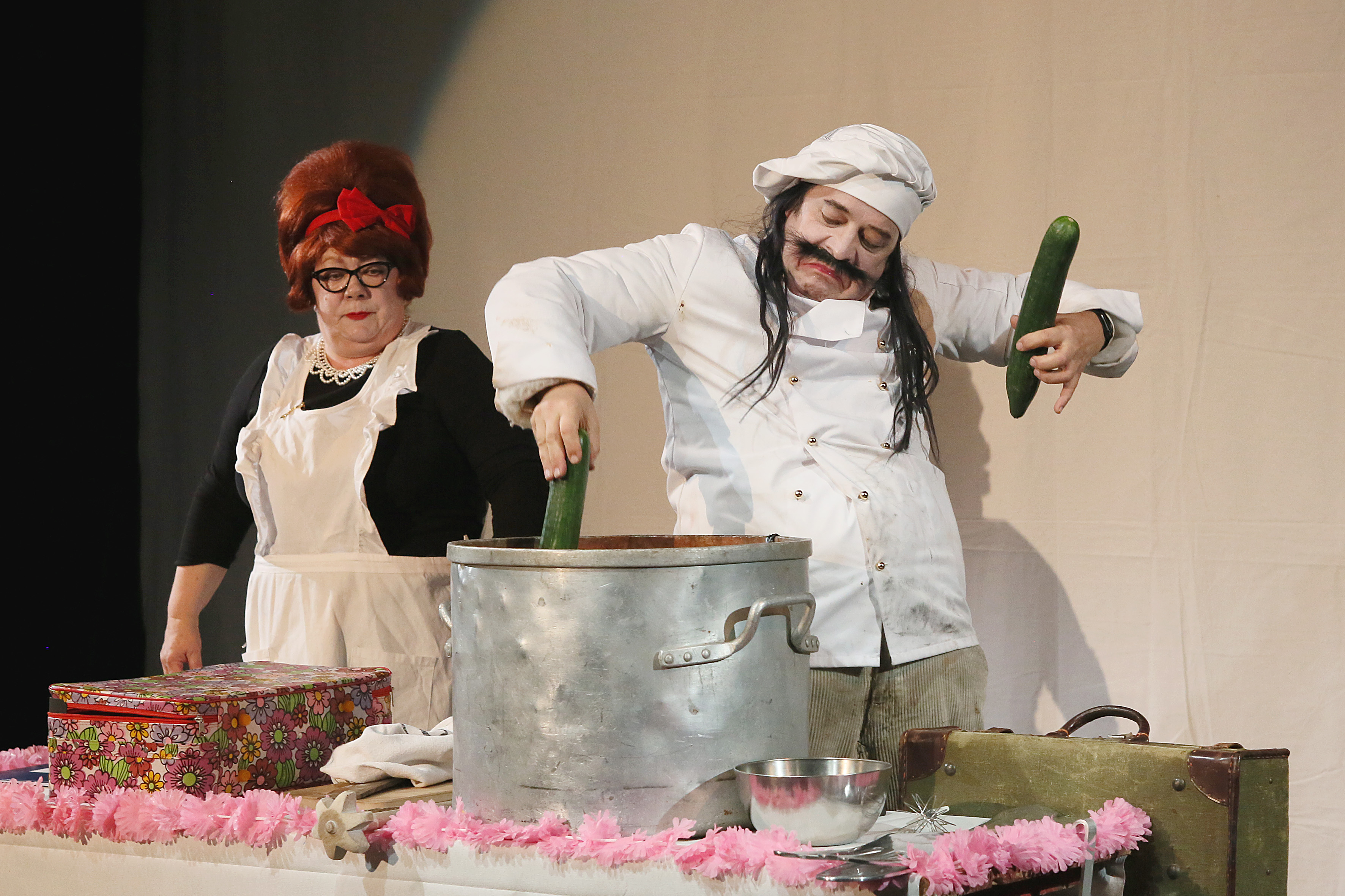 A chubby chef in full cooking gear throws a cucumber into a large pot. He holds another cucumber in his other hand. A woman in a chef's apron stands next to him and watches the action.