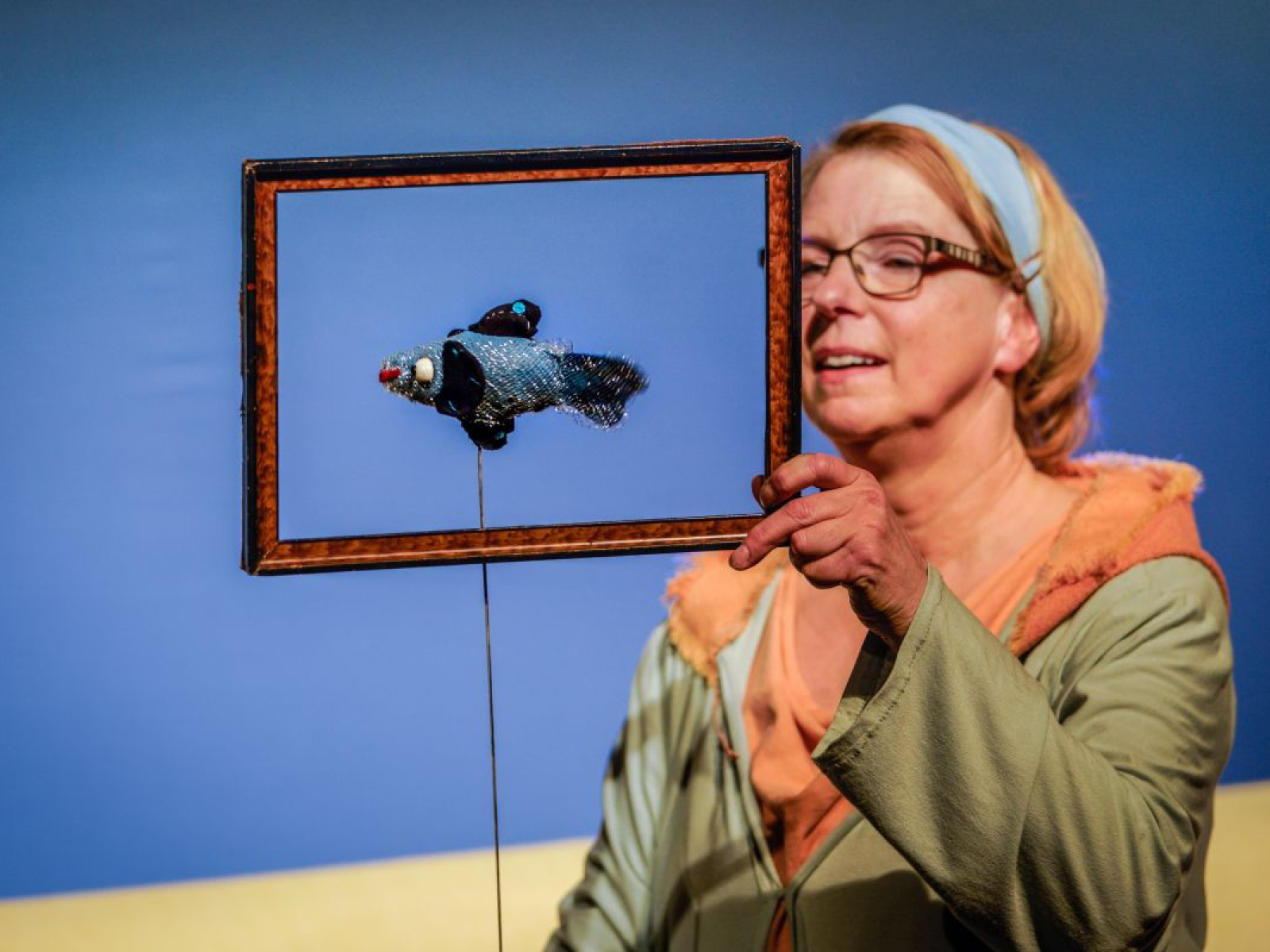 Puppenspielerin Susanne Olbrich hält die Puppe eines kleinen, blauen Fisches mit dunklen Flossen in einen Holzrahmen. Von vorne sieht es aus, als sei der Fisch ein gerahmtes Bild. Sie stehen vor einem blauem Hintergrund.