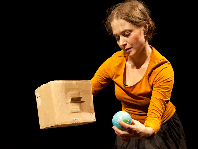 Eine Frau in einem gelben Pullover hält in einer Hand ein Miniaturmodell der Weltkugel und in der anderen einen viereckigen Karton.