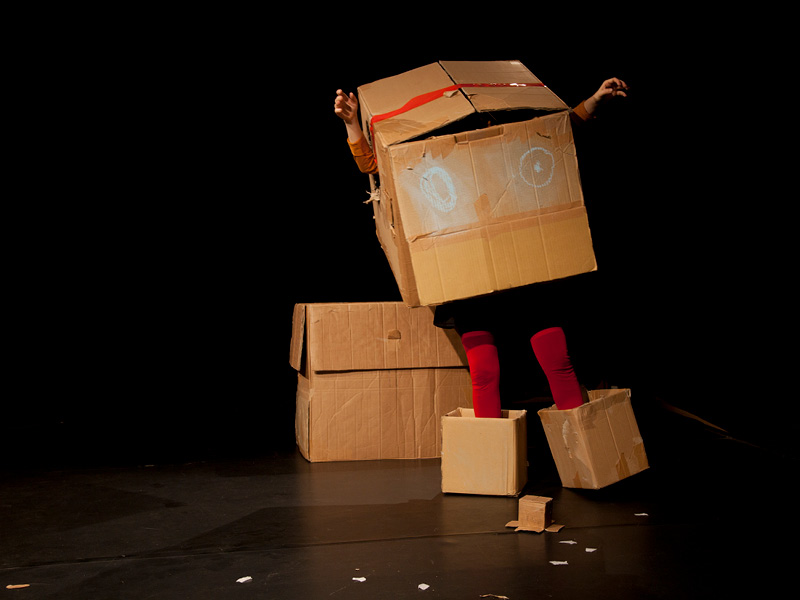 Die Protagonistin steckt in einem großen Karton mit Augen, der ihren Oberkörper verbirgt. Nur ihre, in eine rote Strumpfhose gekleideten Beine schauen heraus und ihre Füße stecken ebenfalls in kleinen Kartons.