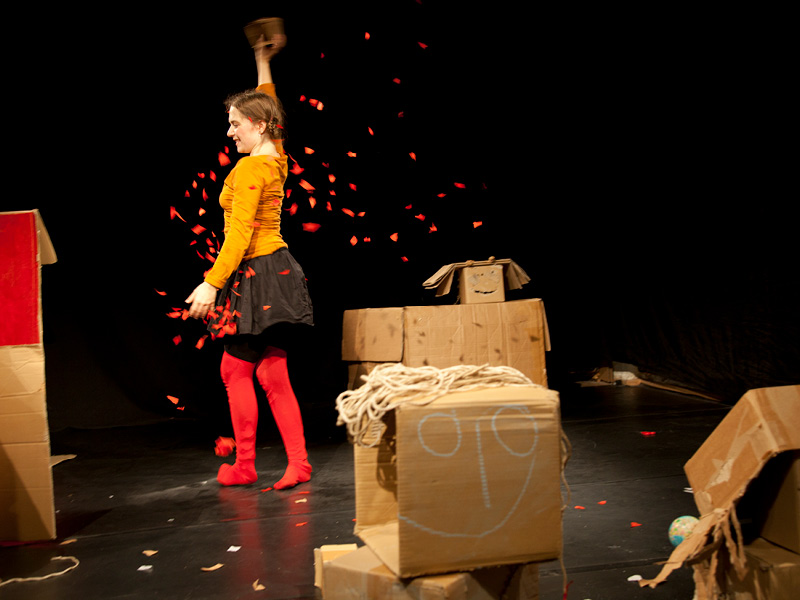 Eine Frau steht, umgeben von Kartons mit aufgemalten Gesichtern und Schnurhaaren, auf der Bühne und lässt rotes Konfetti auf sich regnen.