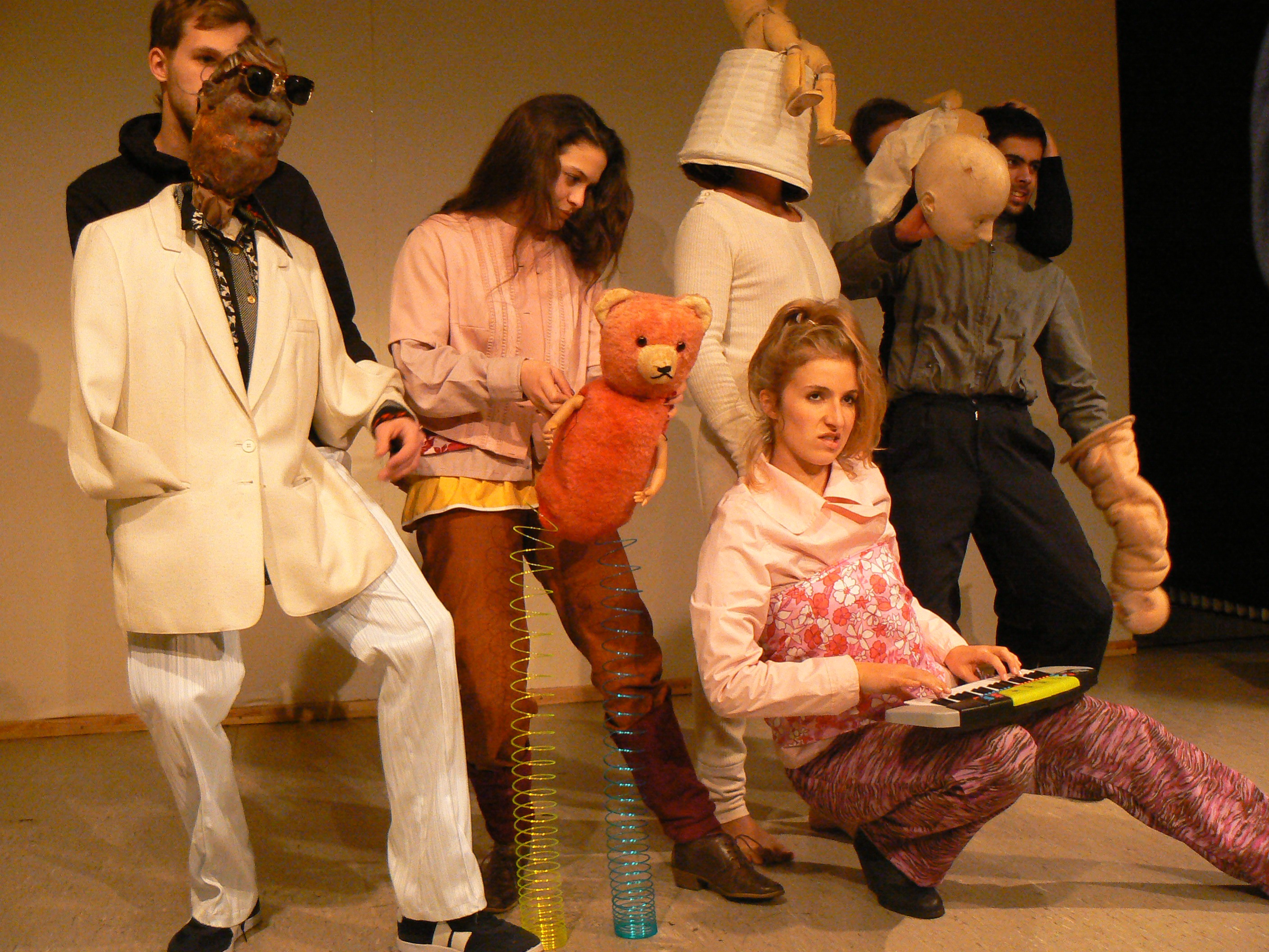 Fünf StudentInnen in bunten, eigenwilligen Klamotten bewegen und lenken verschiedene Puppen. Beispielsweise einen Teddibär mit Sprungfederbeinen, Puppenköpfe, einen Torso in weißem Jackett und Kartoffelähnlichem Kopf mit Sonnenbrille.