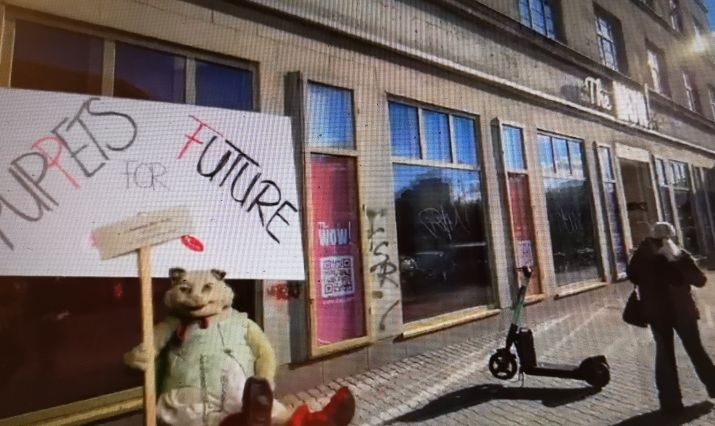 Ein Katze mit Overall hält ein Schild in die Höhe. Hinter ihr ist ein Schild mit dem Namen Puppets for Future angebracht. Sie steht auf einem Gehweg vor den Fenstern der Schaubude.