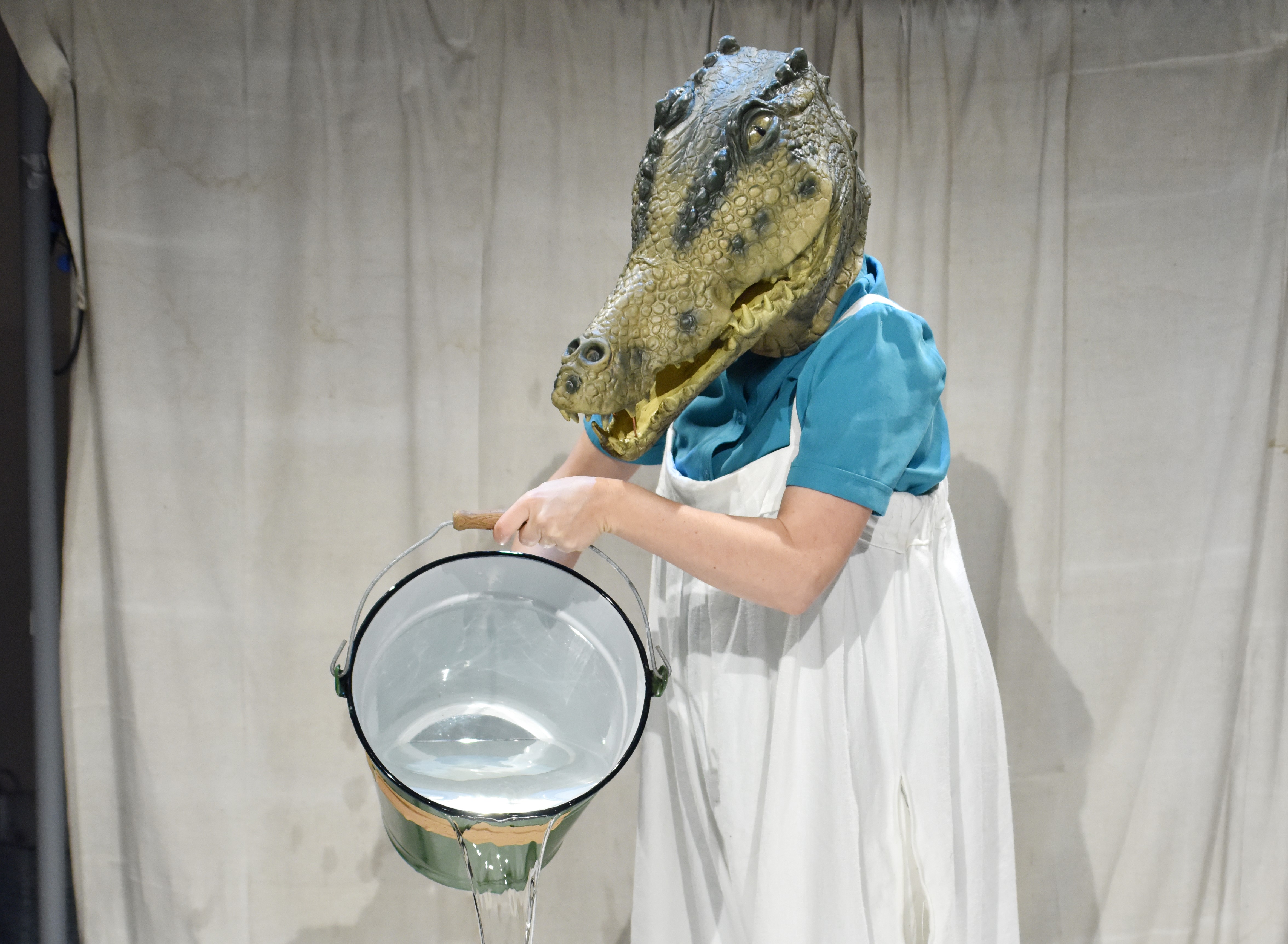 Puppenspielerin Ulrike Kley trägt eine echt aussehende Maske eines Krokodils mit spitzen Zähnen und langer Schnauze auf dem Kopf und schüttet einen Eimer voll Wasser auf die Bühne.