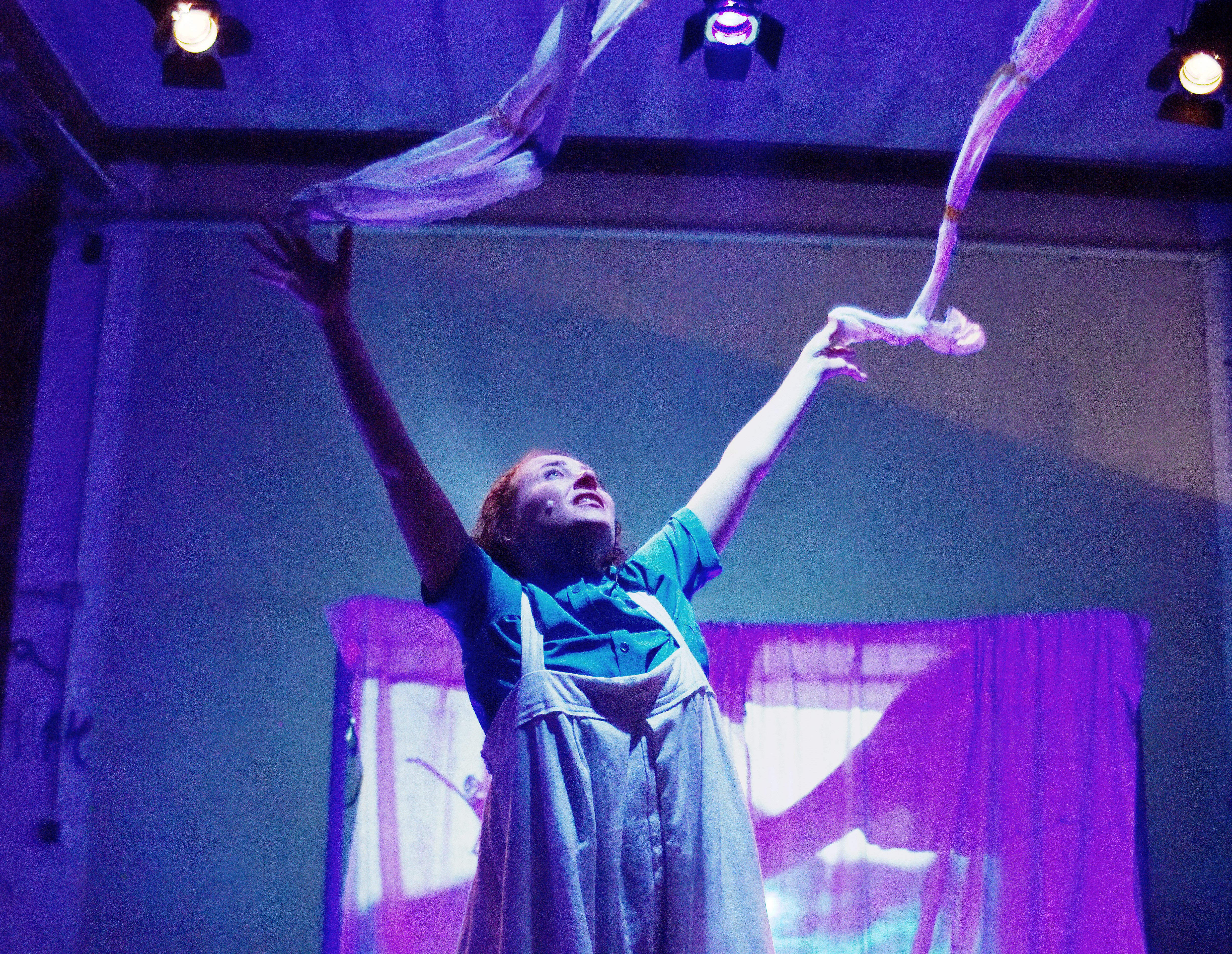 Ulrike Kley wirft mit ausgestreckten Armen zwei lange, weiße Bänder in die Luft. Der Raum hinter ihr zeigt eine Projektion und ist blau, magentafarben beleuchtet.