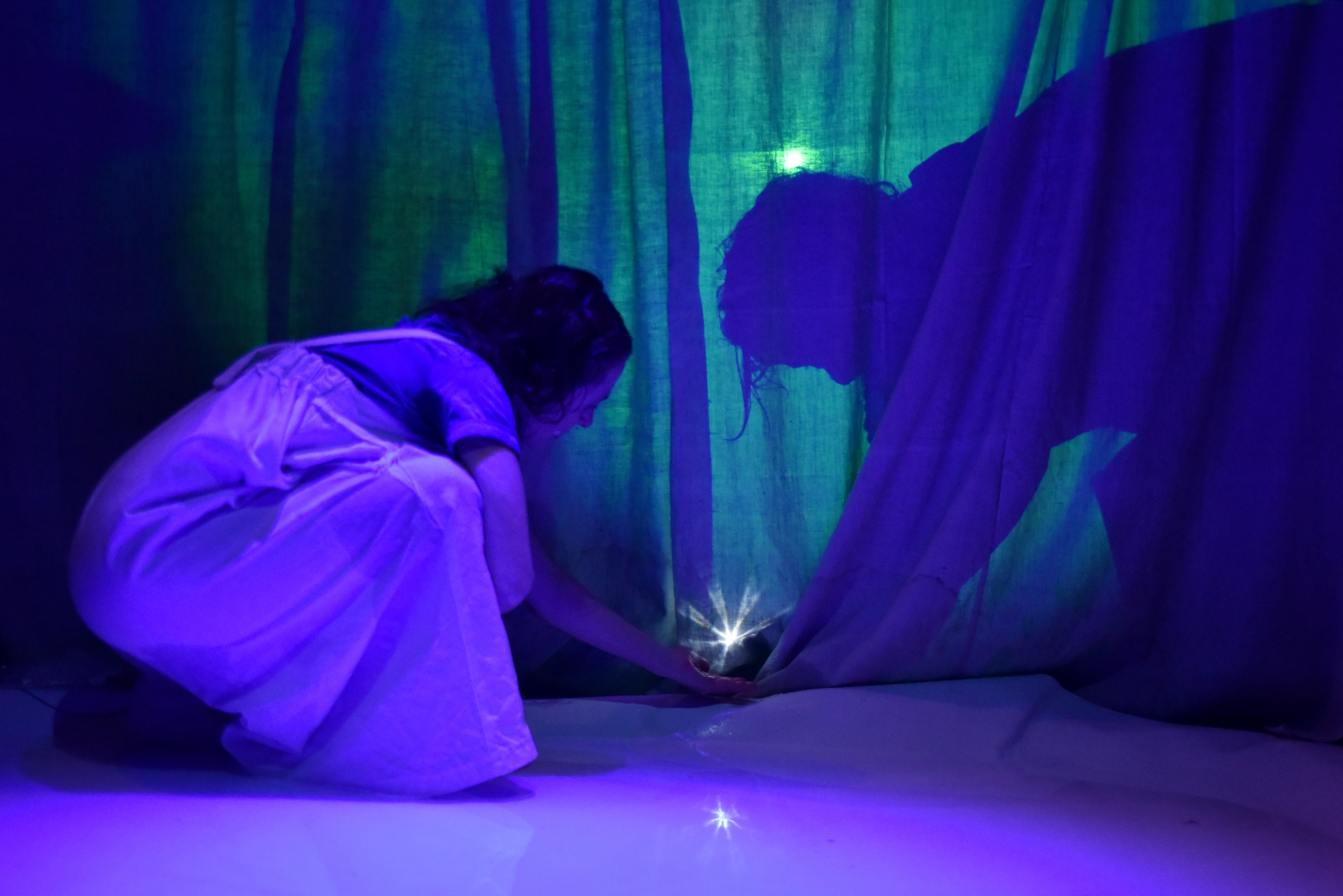 Samira Wenzel hält ein kleines helles Licht in der Hand, das sich in der nassen Bühnenoberfläche einspiegelt. Sie ist umgeben von diffusem, blau, grünem Licht und zarten Vorhängen.