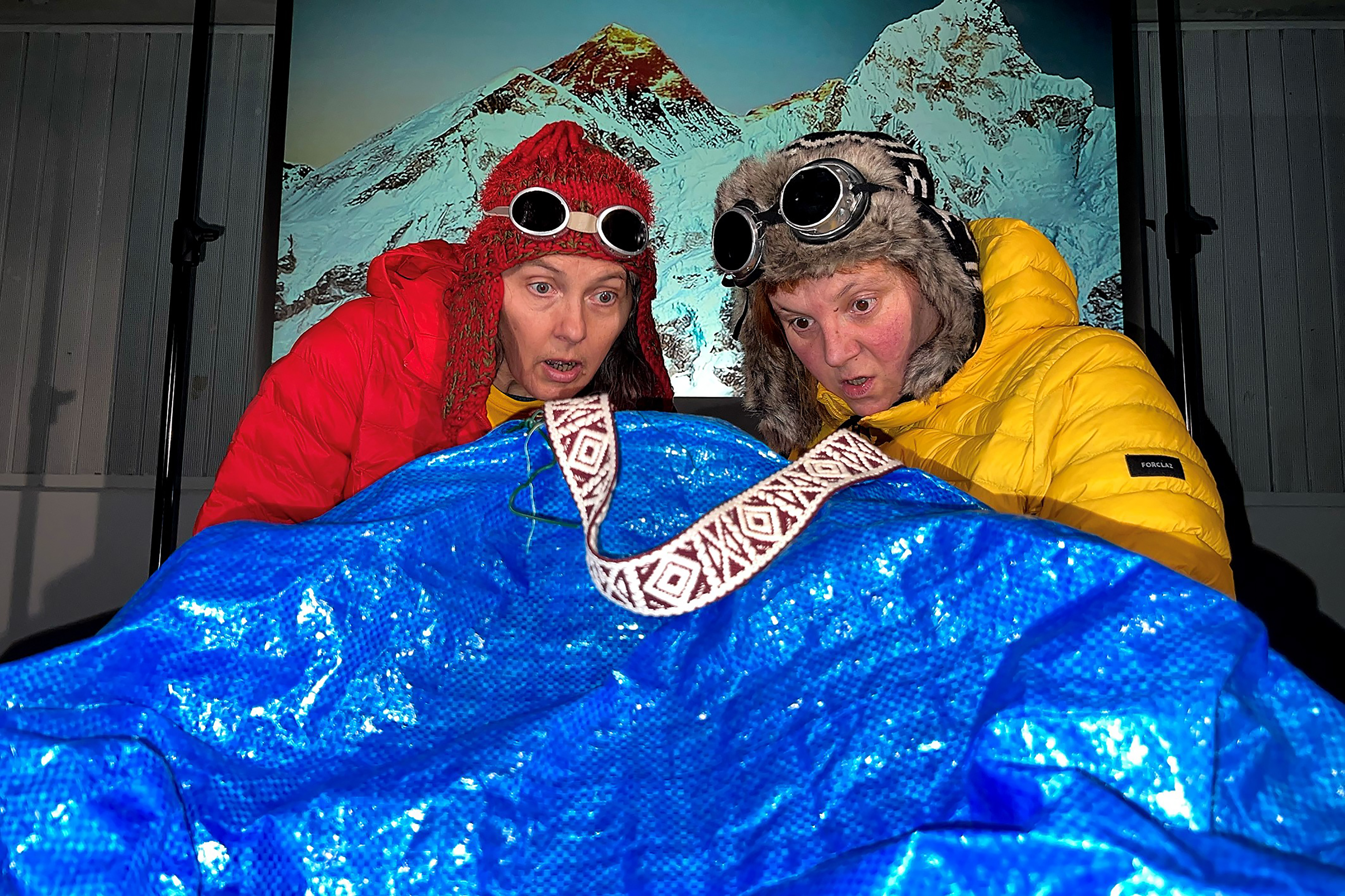 Alexandra und Eva Kaufmann haben ihre Sonnenbrillen über ihre dicken Ohrenwintermützen gezogen und schauen entgeistert in eine blaue Ikeatasche. Der Inhalt ist verborgen. Im Hintergrund ist ein Aufsteller des Mount Everest zu sehen.