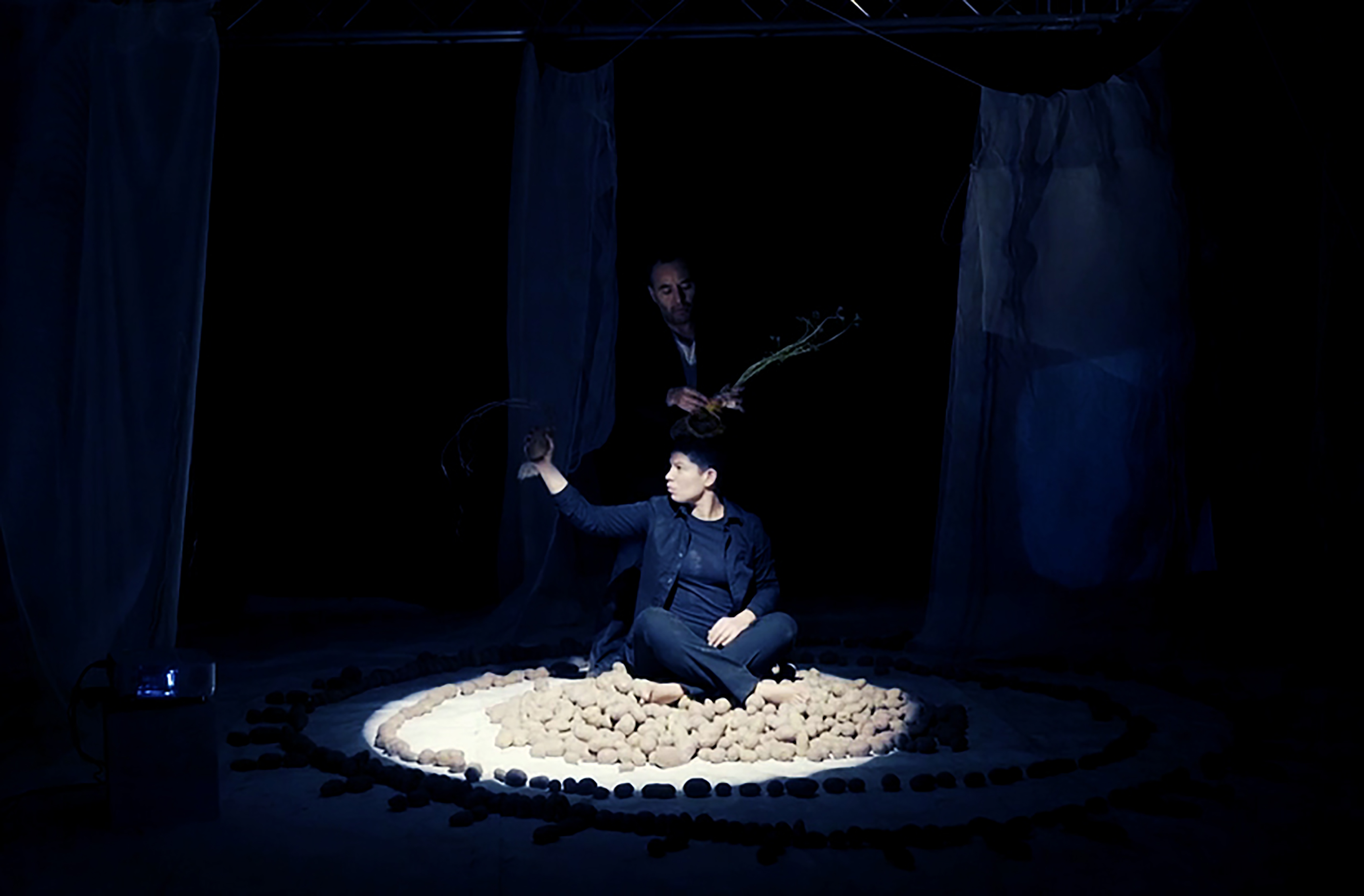 Performerin sitzt auf erdigen Kartoffeln mit erhobener Hand im Schneidersitz in einem hell erleuchteten Kreis. Im Hintergrund ist ein zweiter Performer.