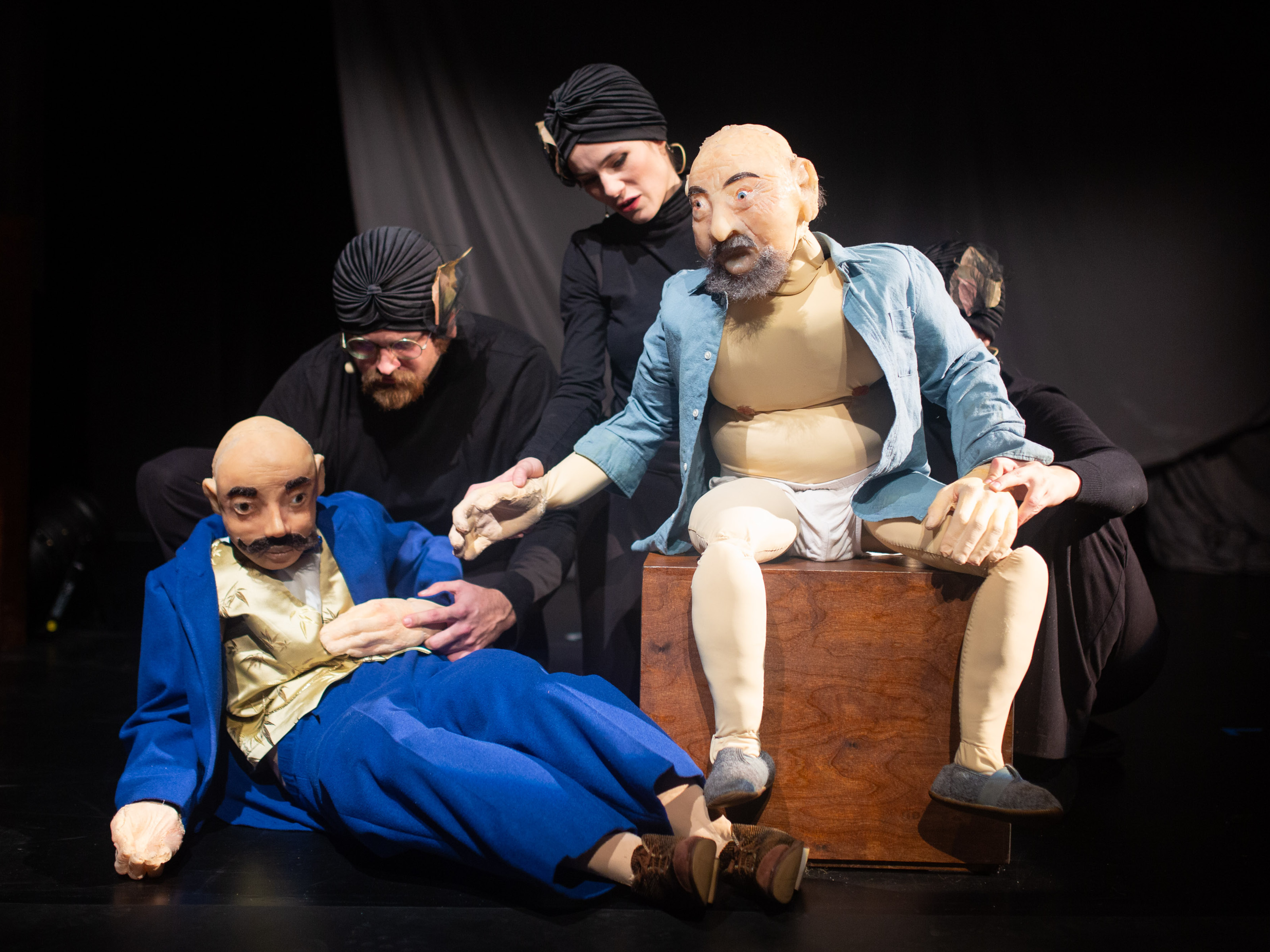 The puppet of Prince Sternenhoch is dressed in high quality blue suit and golden vest next to the puppet of an older man with buttoned shirt and white underpants. Behind them are the puppeteers in black clothes with black hoods.