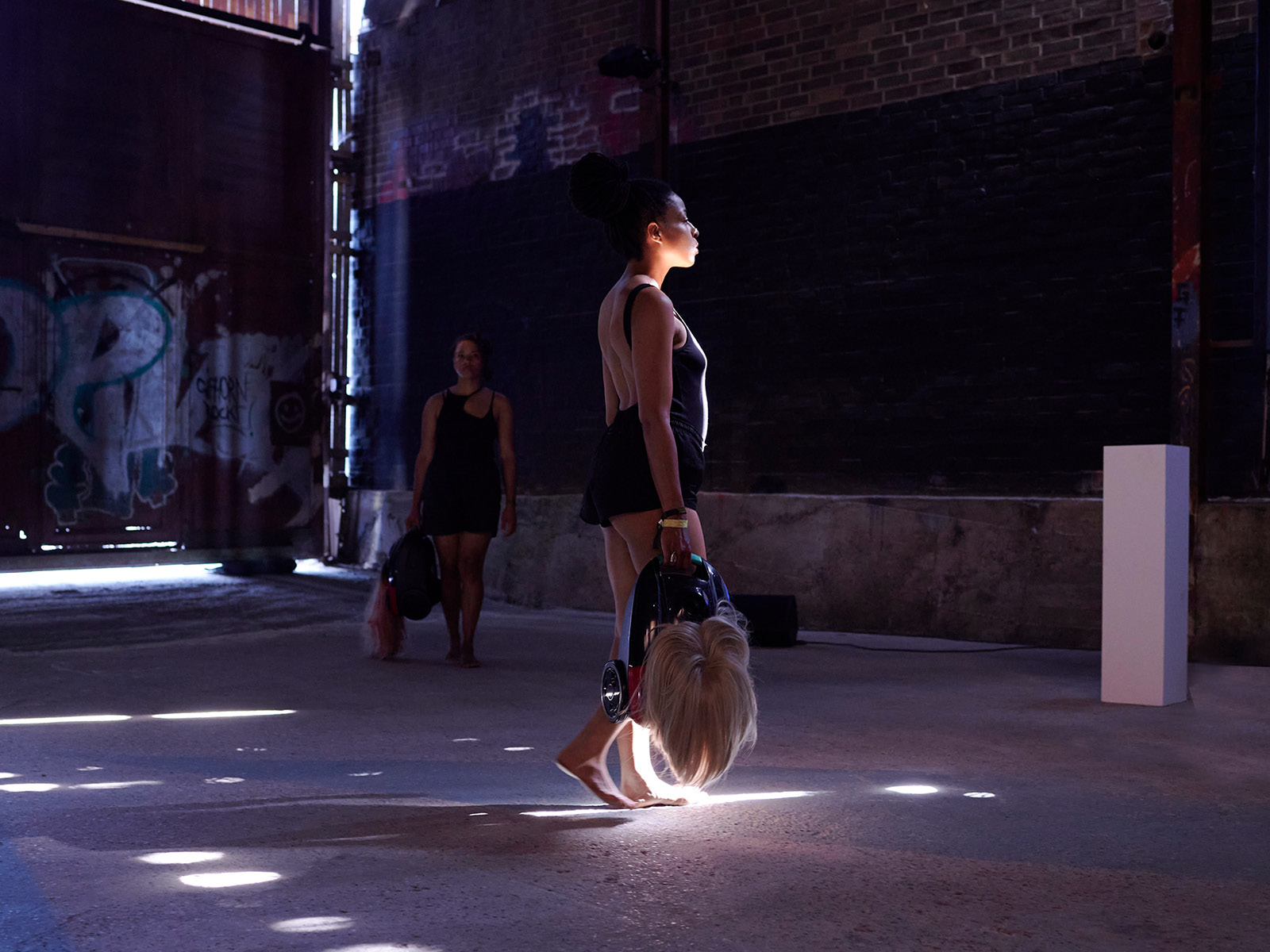 Two women in black bodies wander through a room into which isolated rays of sunlight shine.
