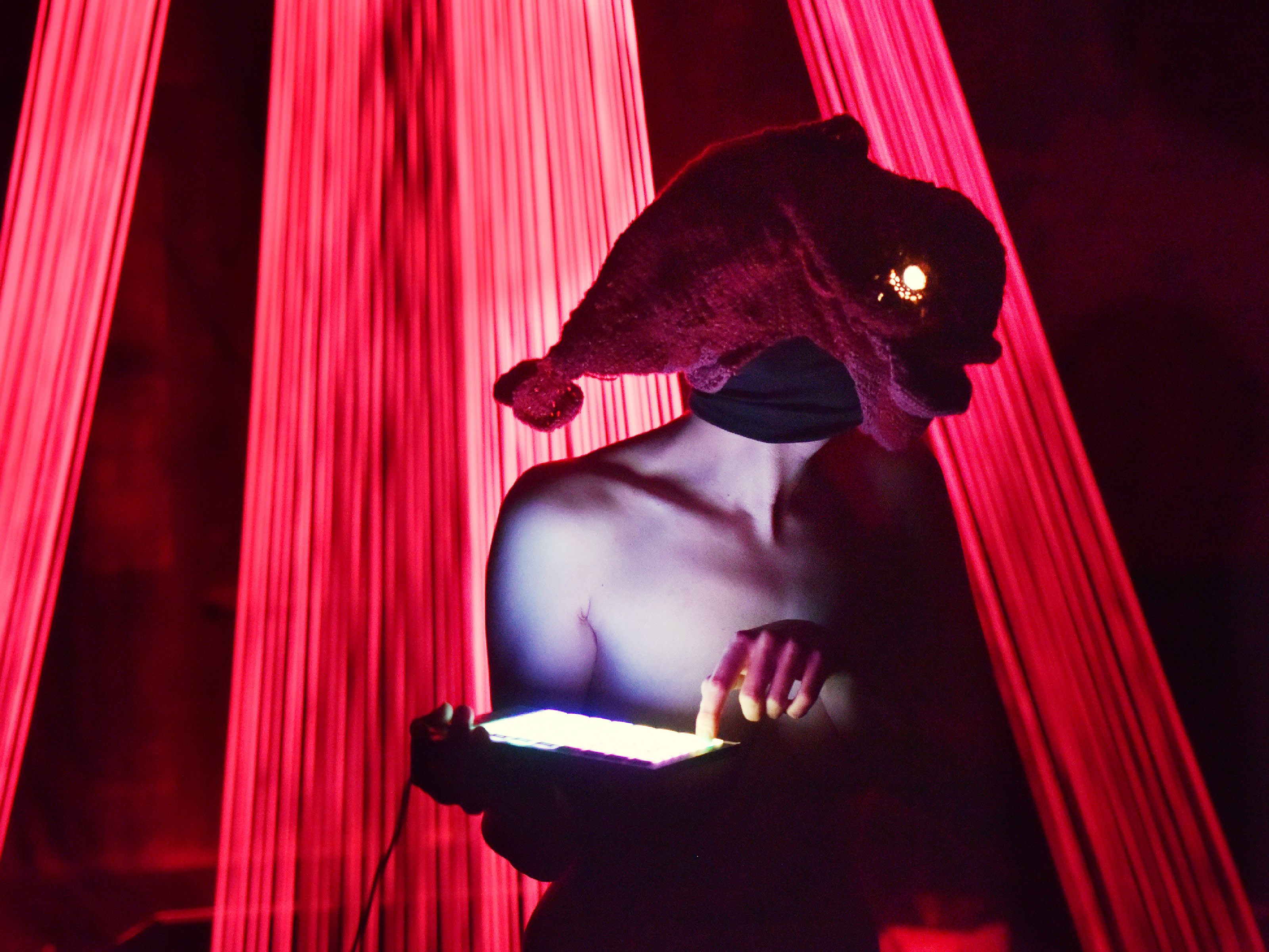 A naked performer wears a crocheted fish on her head, which has a glowing lamp eye. Her face is completely covered by the fish and a black mask. She presses down one of the luminous buttons of an electronic device.