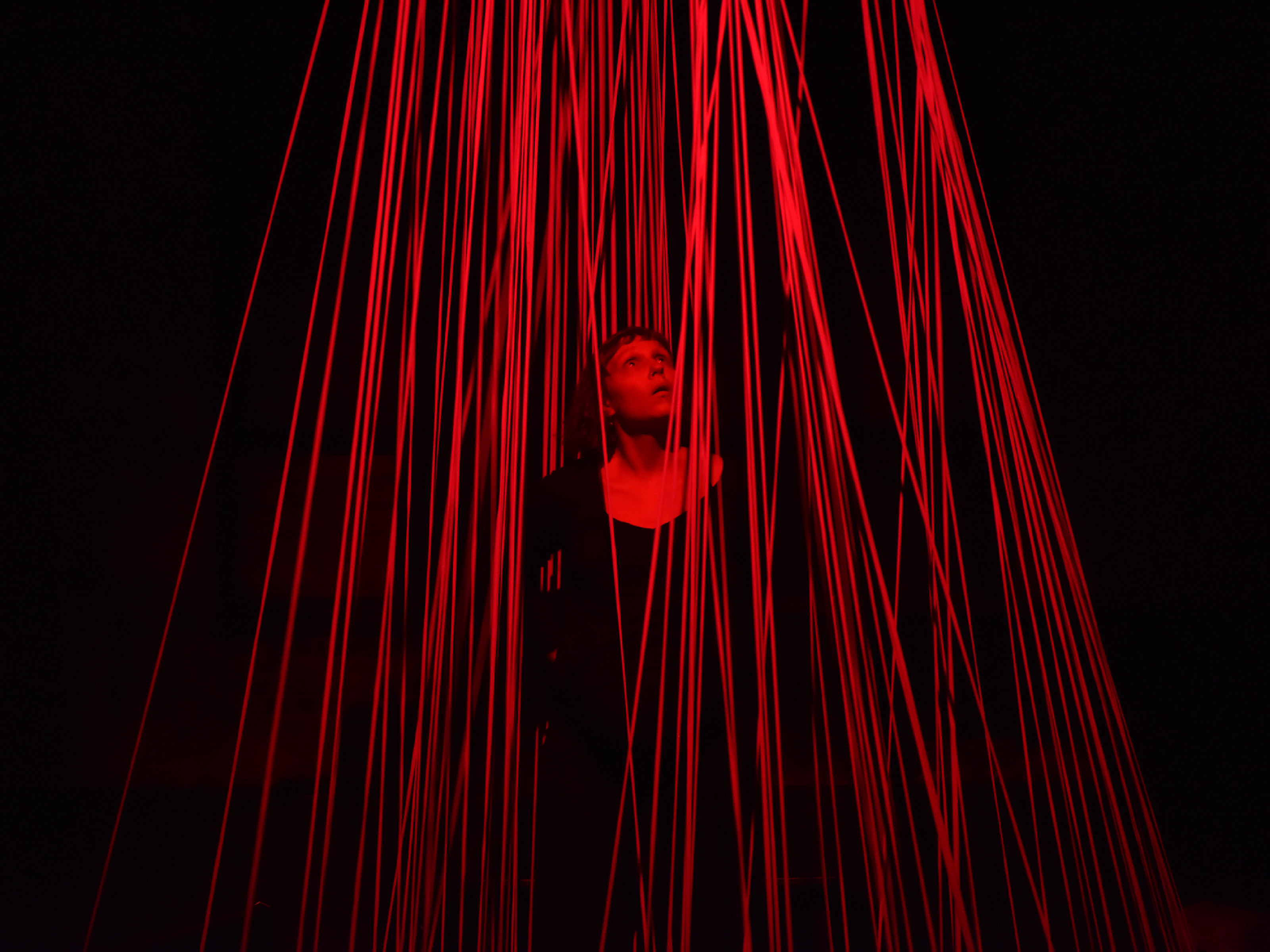 A performer dressed in black stands between numerous thick cords leading from the floor to the ceiling with an astonished expression on her face and her eyes fixed on the ceiling. The entire scene is bathed in red light.