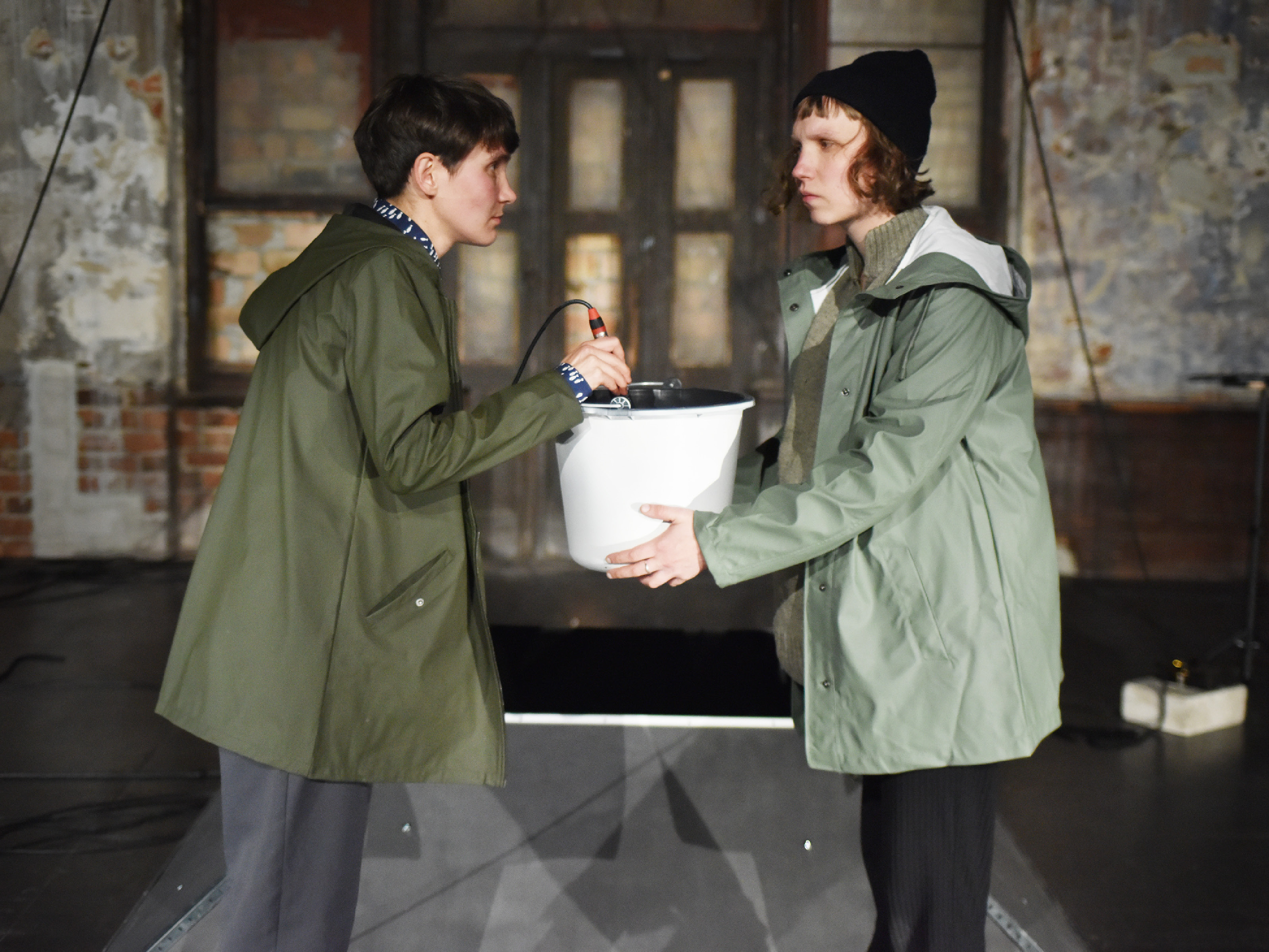 Two performers in green rain jackets face each other. One holds a white metal bucket in her hand, while the other holds an electronic device in the bucket.
