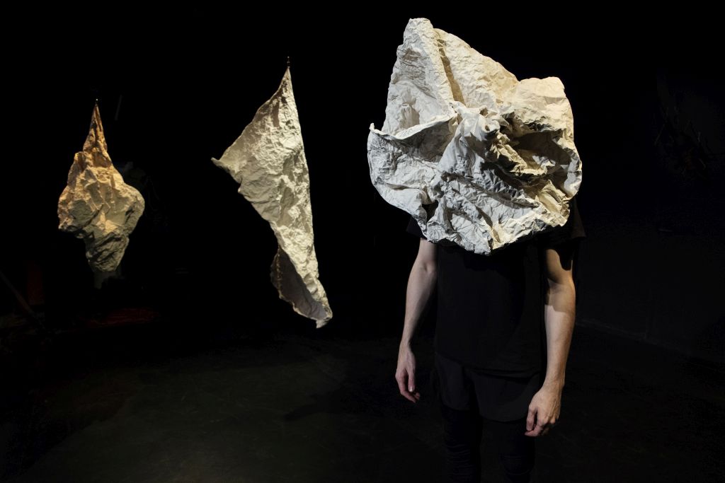 Performer mit papierähnlichem Material über dem Kopf; von ihm sind nur die Arme und schwarze Kleidung sichtbar. Im Hintergrund sind zwei von der Decke hängende weitere papierähnliche Formen zu sehen.