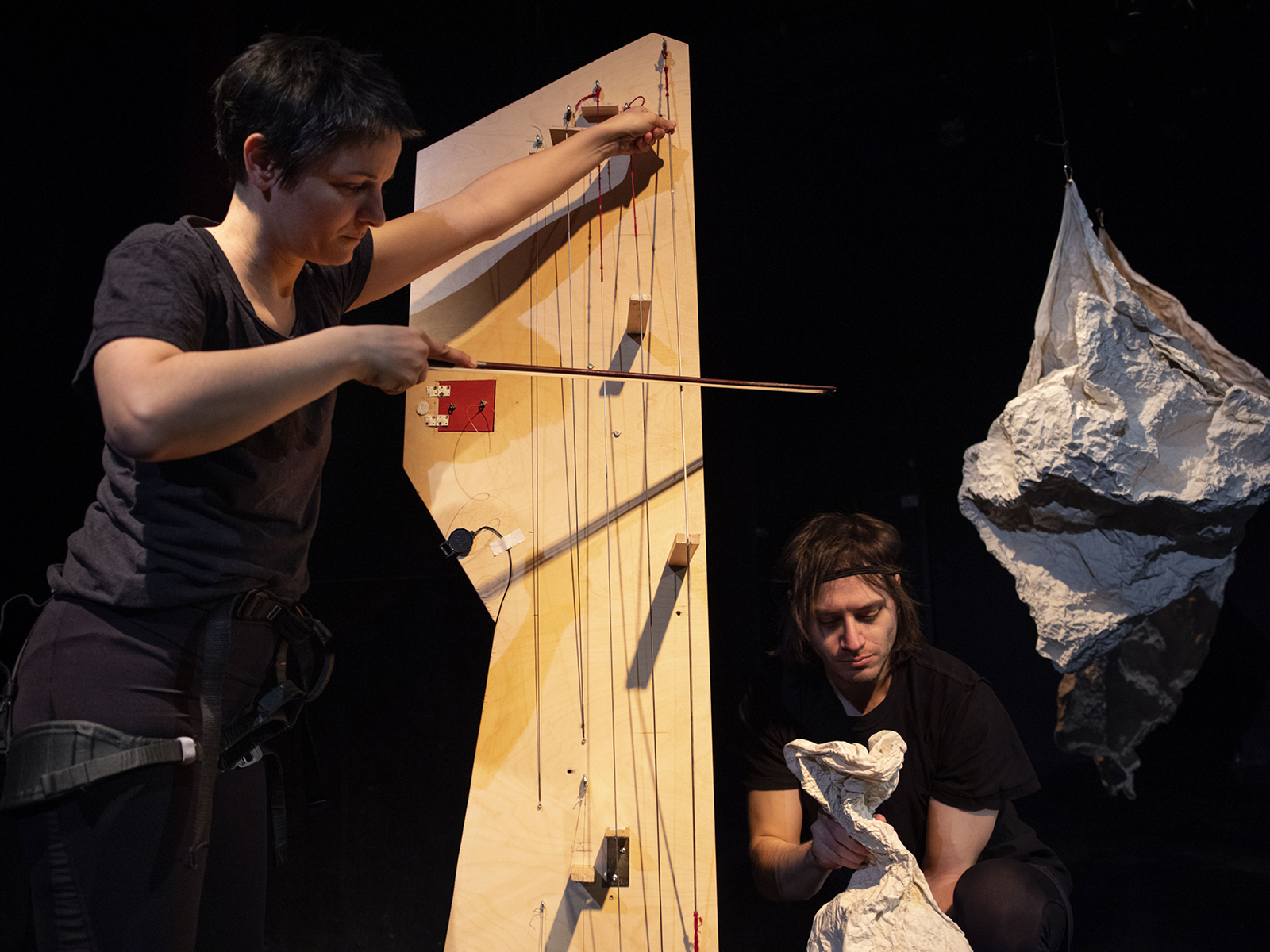 Performerin steht und streicht mit Geigenbogen die Saiten eines selbstgebauten Holzinstruments. Rechts daneben hockt ein weiterer Spieler, der mit den Händen ein papierartiges Material animiert als ob es eine Puppe wäre.
