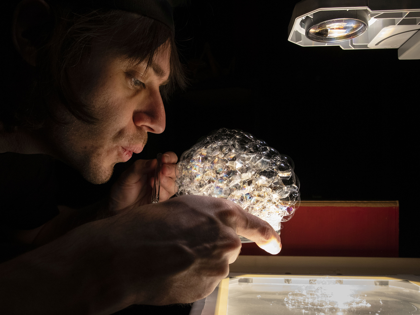 Ein Performer bläst mit einem Strohhalm Schaumblasen in einer Glasschale, die von einem Overhead-Projektor beleuchtet wird.