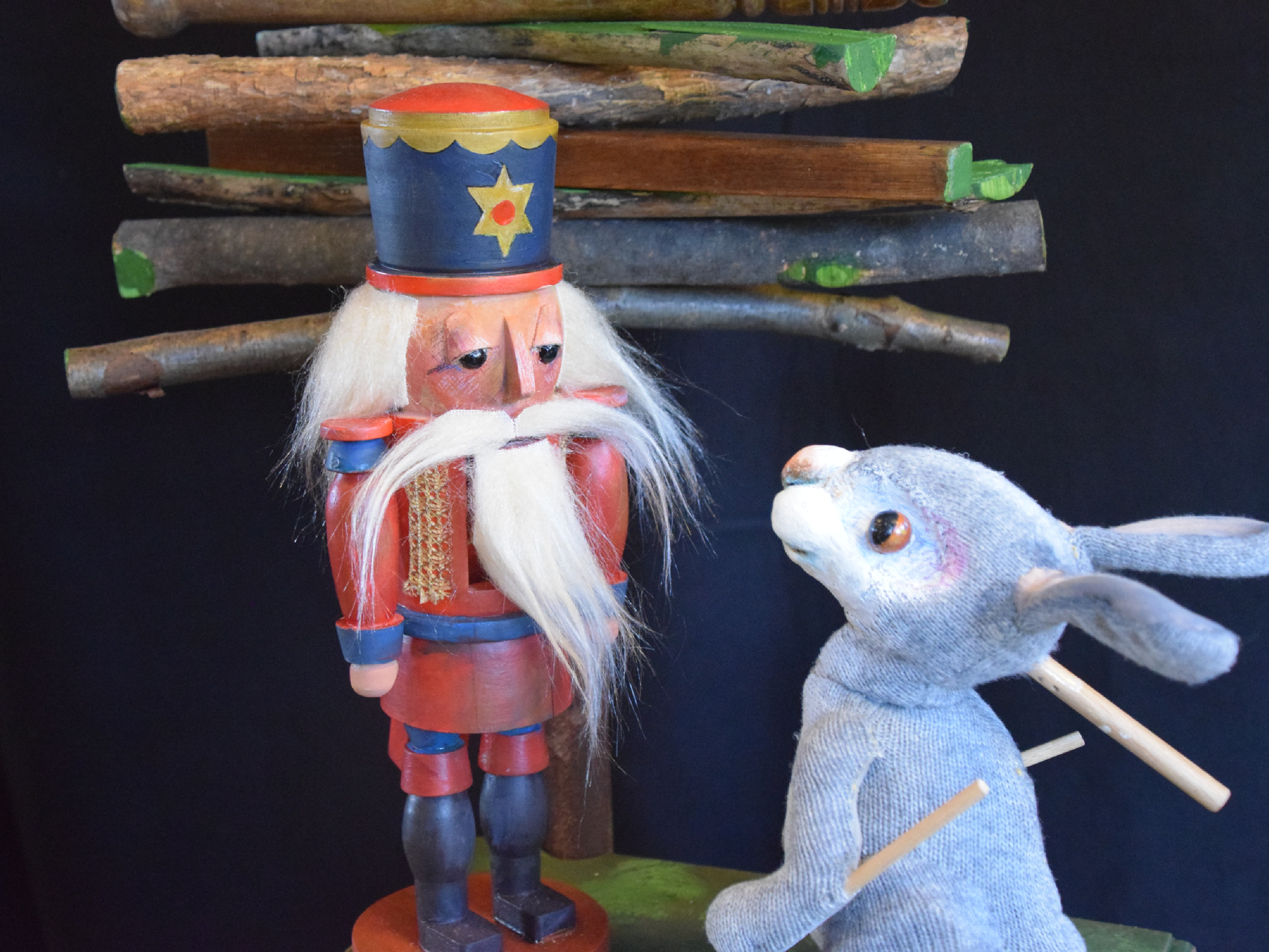 The rag doll of the little rabbit stands in front of a nutcracker with white hair and long bar. In the background there is a Christmas tree made of plain, leafless branches.