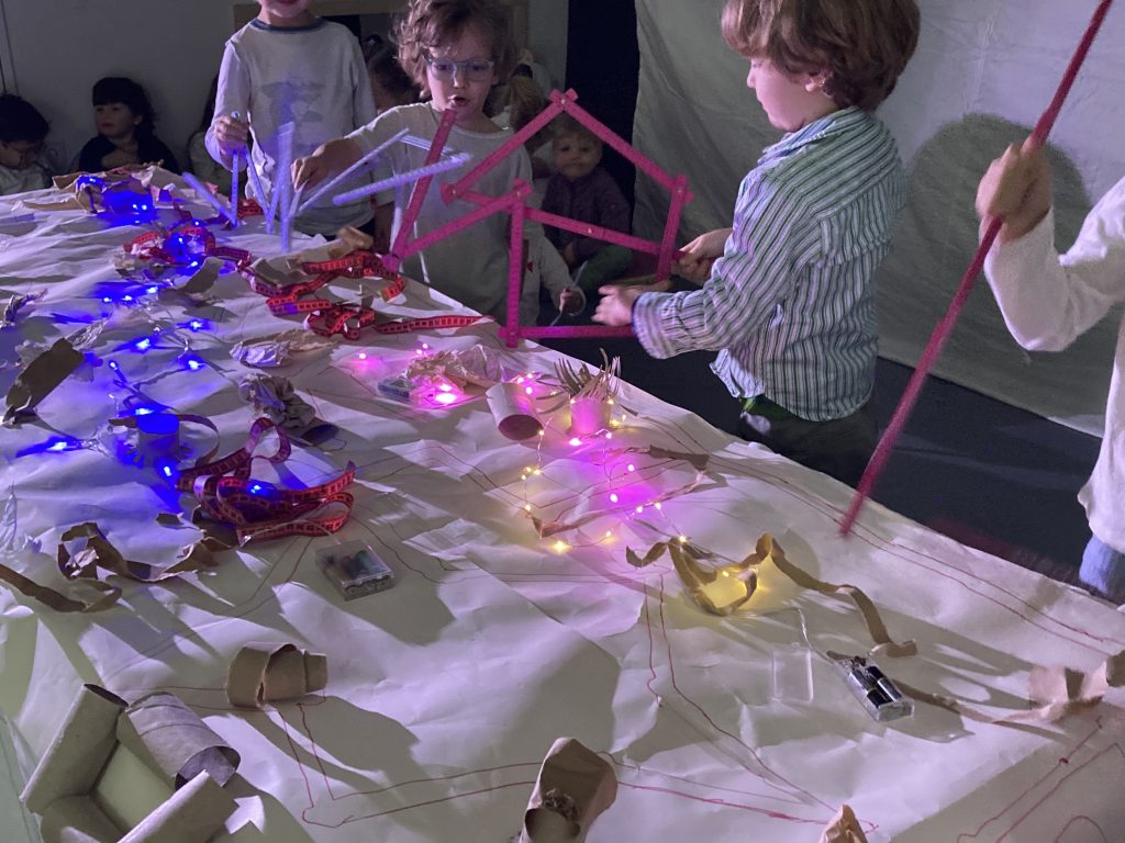 Um einen Tisch, der mitLichterketten nd papierobjekten gestückt ist, stehen 3 Kinder, die mit geformten Zollstöcken zwischen den Tish-Objekten staksen. die Zollstöcke erinnern teilweise an Beine von Tieren