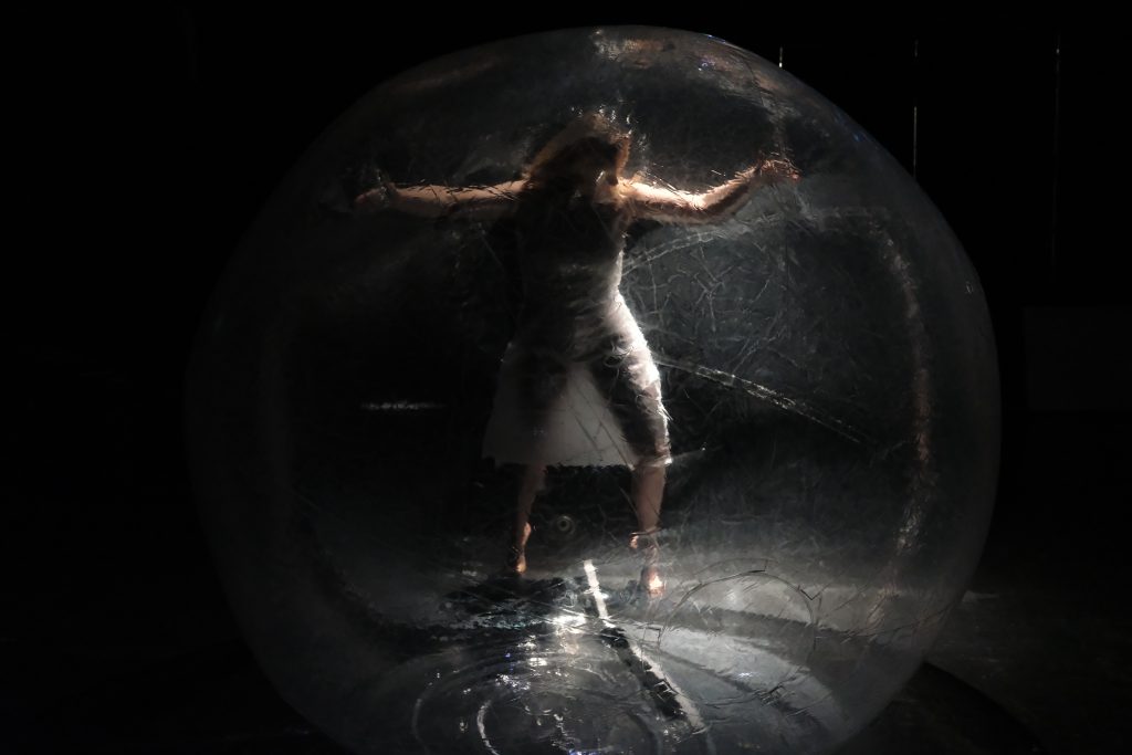 Eine Person ist in einem transparenten Ball. Sie trägt weiße Klamotten und ist von einem harten Licht von rechts beleuchtet.