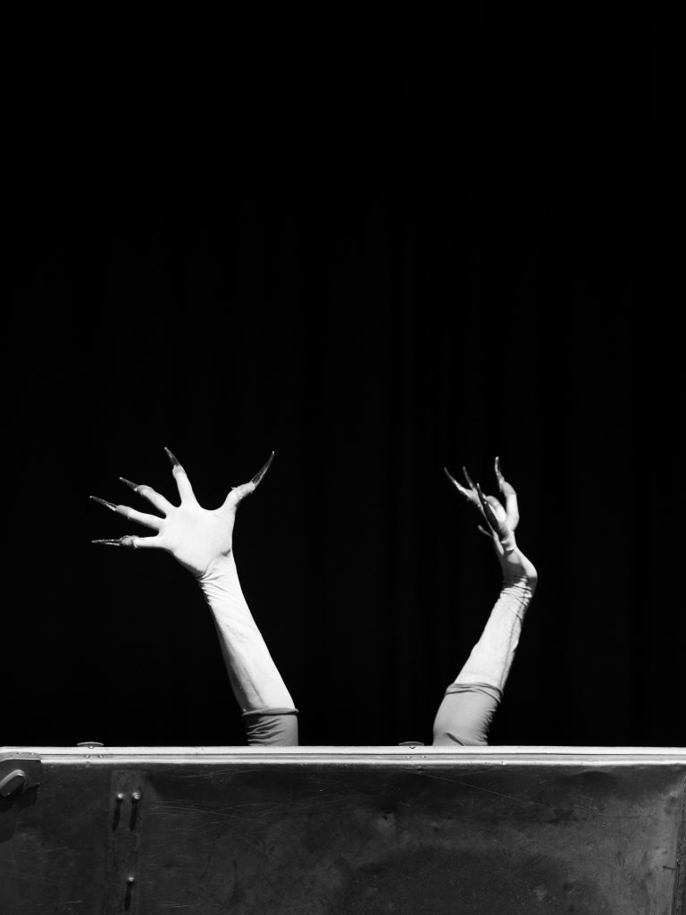 Überstreckte Arme in weißen Handschuhen recken sich aus einem Sarg.