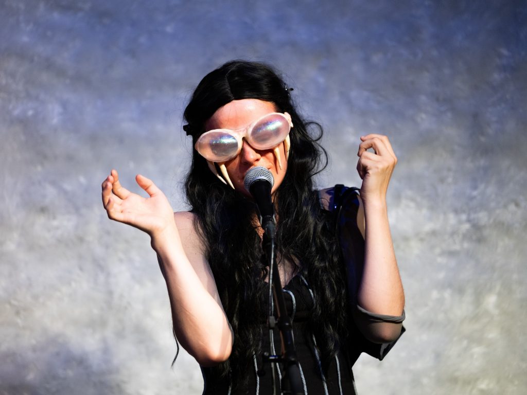 Eine junge Frau mit schwarzer Perücke trägt eine Brille mit Fangzähnen. Sie steht vor einem Mikrofon und singt