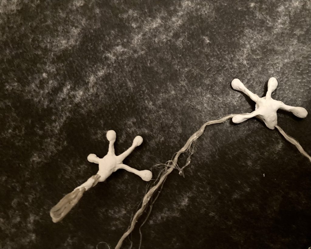 Zwei reptilienartige Hände aus weißer Knetmasse liegen nebeneinander auf einem dunklen Hintergrund