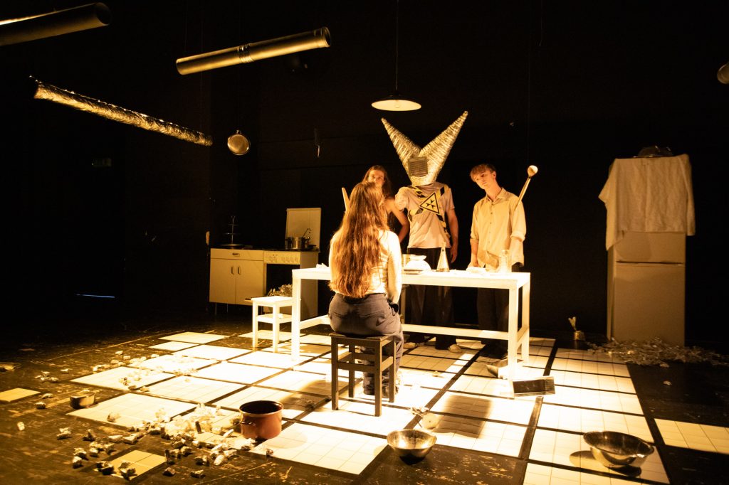 Drei Personen sitzen und stehen in einem Bühnenküchenraum.