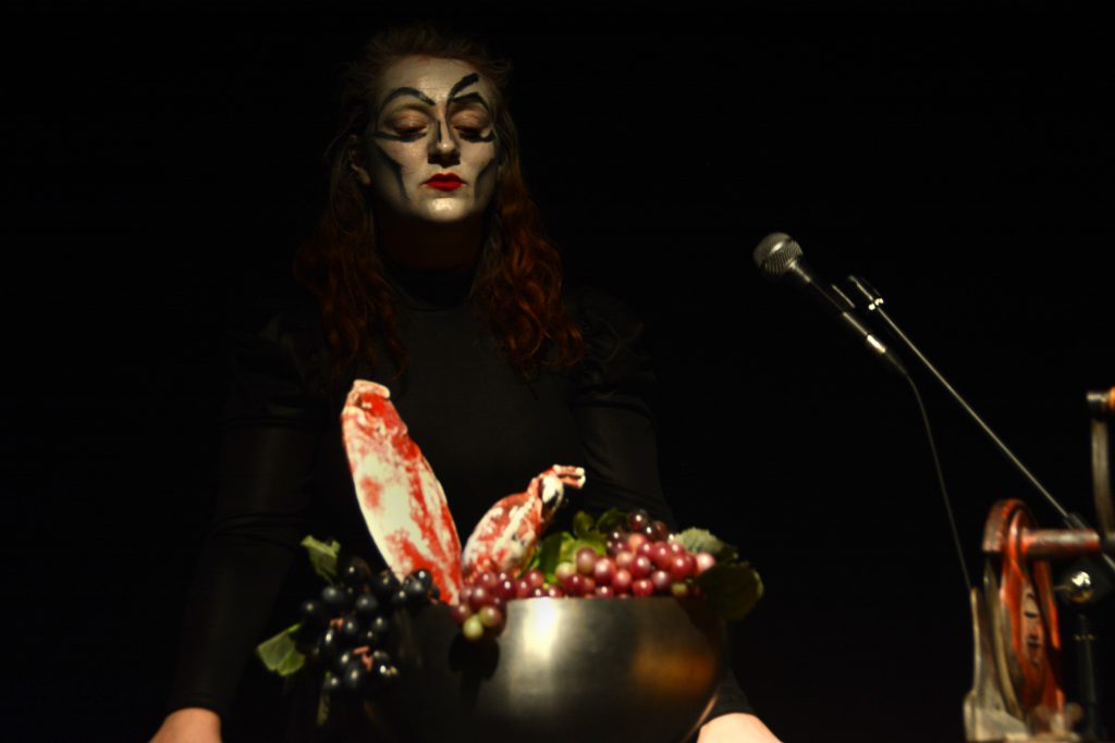 Eine Person mit silbern geschminktem Gesicht hat vor sich einen Topf mit Obst