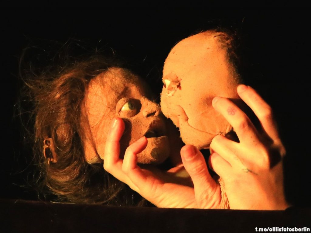 Zwei Puppengesichter aus Stoff werden von zwei menschlichen Händen nahe aneinander gehalten und schauen sich in die Augen.