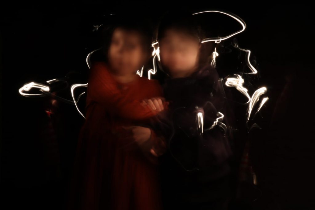 vor schwarzem Hintergrund sind schemenhalt zwei Kinder erkennbar, deren Gesicht in der Bewegung verschwommen ist, um die Köpfe herum gibt es Lichtfäden, die an ein Geflecht erinnern