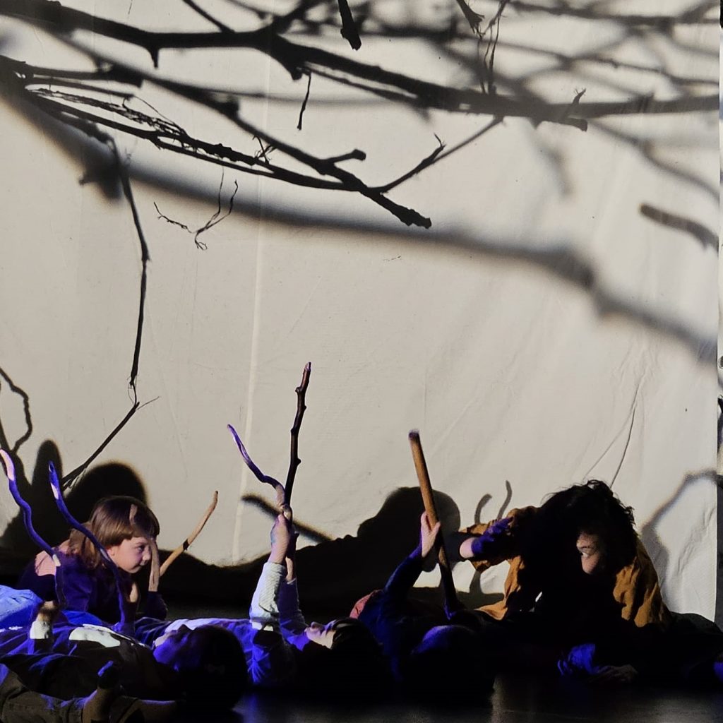 vor einer hellen Wand sieht man projizierte und echte Birkenzweige, die mittels eines Overheadprojektors an die Wand geworfen werden. Am Boden liegen Kinder, die Stöcke vor ihren Köpfen hochhalten