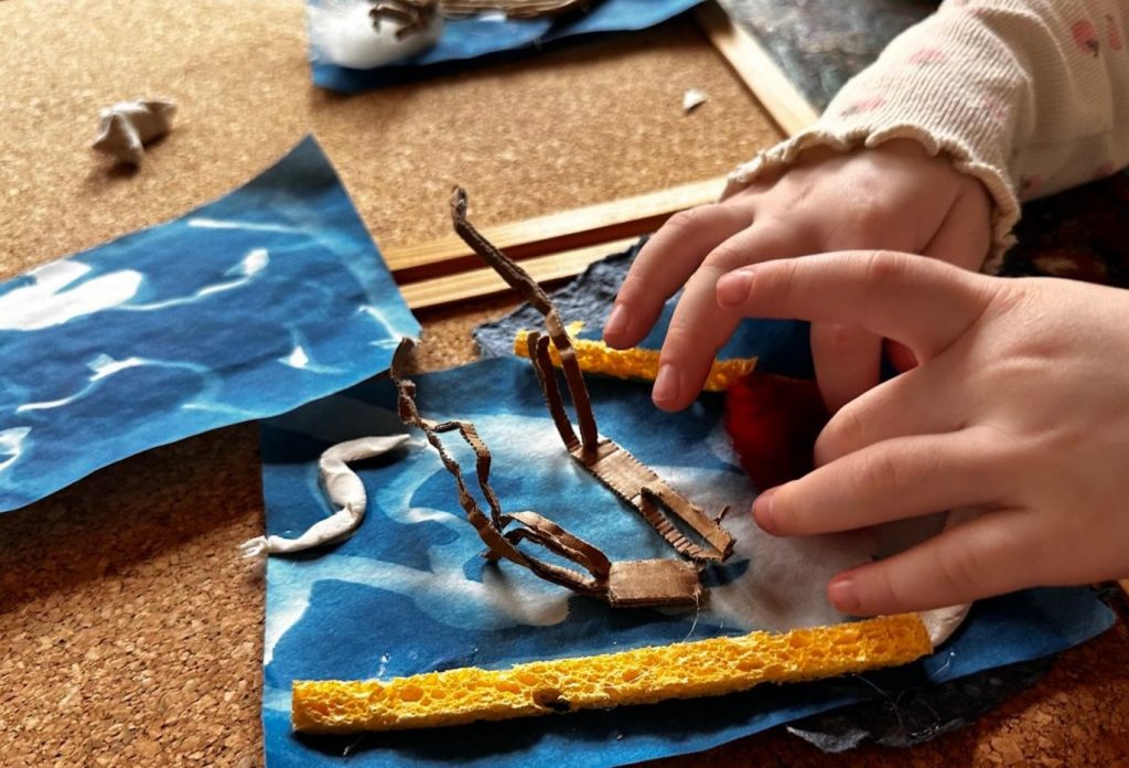 auf einem beigen Untergrund liegen blau belichtete Fotopapiere, auf denen kleine Wurzeln und ein längliches Schwämmchen liegen. Zwei hände ertasten die Objekte