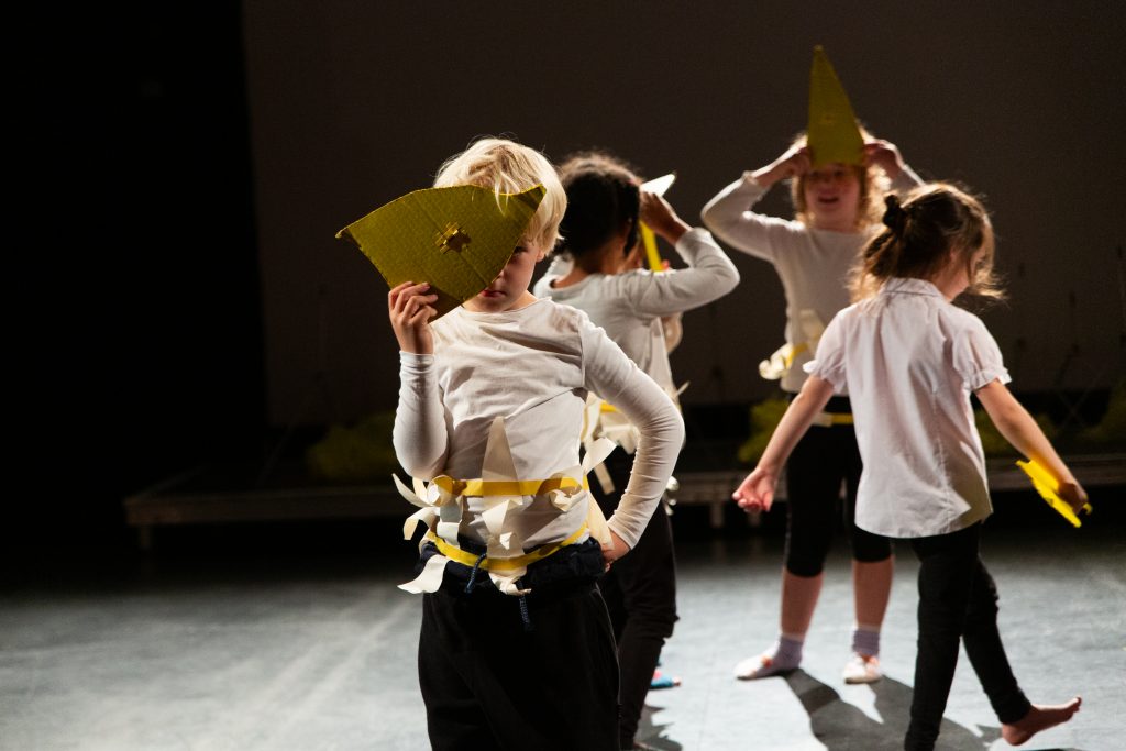 Auf einer Bühne mit schwarzem hintergrund laufen 4 Kinder mit weißen oberteilen und gelben Schablonen über die Bühne. Ein Kind im Vordergrund hält sich die Pappschablone wie eine Maske vor das Gesicht, so dass nur ein Auge etwas hervorschaut.