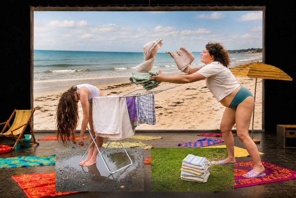 Beach scene with a clothesline displaying various fabrics and textile objects. A person stands to the right of the line with raised arms, while the sea and sandy shore are visible in the background.