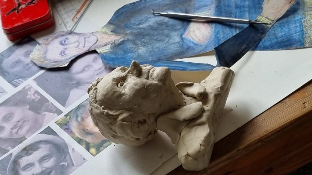 Alt Text (English): A small clay sculpture of a human head with closed eyes rests on a worktable. Additional sculpted clay pieces are placed beside it, while a blue-toned reference image or artwork lies in the background. The scene suggests an artist’s workspace or studio during the process of creating a ceramic or clay figure.