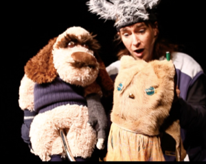 A costumed performer stands against a black background holding two hand puppets. On the left is a brown-and-white dog puppet wearing a dark vest, and on the right is a light-colored character puppet with blue eyes and a long dress. The scene is warmly lit and shown in close-up.