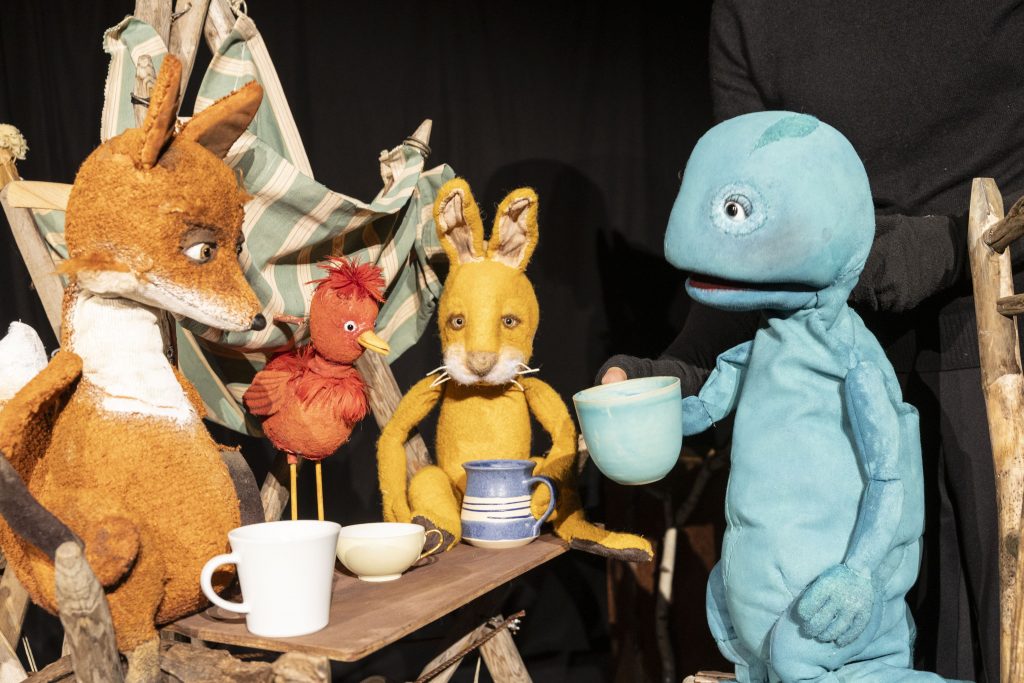 Several animal and character puppets are gathered around a small table with cups. A yellow rabbit puppet is positioned in the center, while a blue character puppet on the right holds a cup. Additional puppets are partially visible on the left. The scene is set against a dark background.