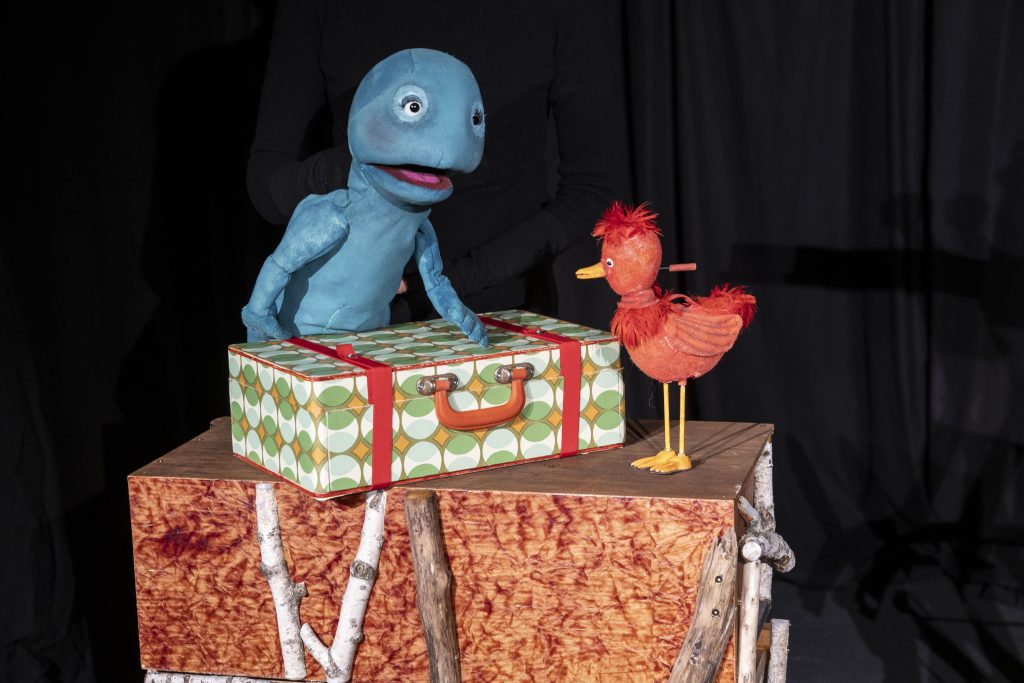 A blue character puppet and a red bird puppet stand on a wooden stage surface against a black background. A patterned suitcase with an orange handle is positioned between them. Parts of a puppeteer dressed in black are visible in the background.