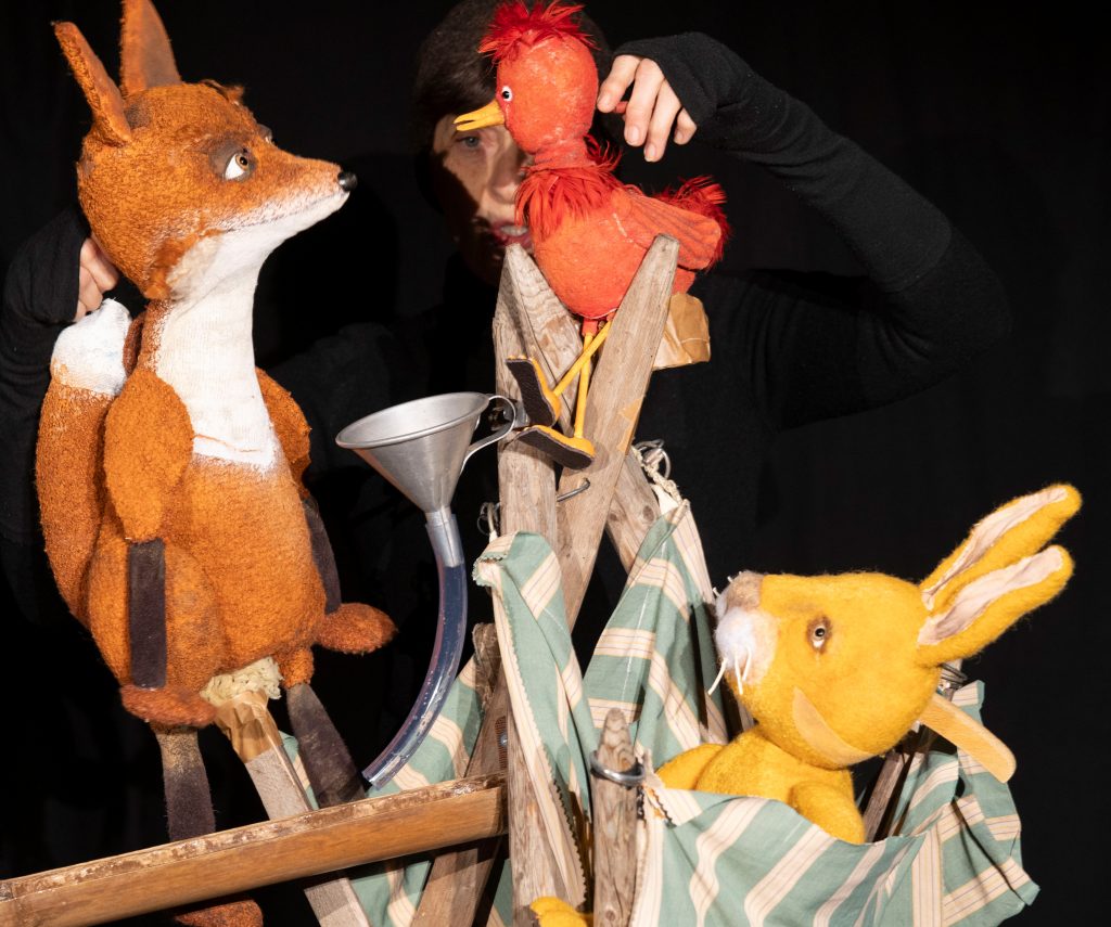 A person dressed in black operates several animal puppets on a stage against a black background. A fox puppet stands on the left beside a wooden structure holding a red bird. On the right, a yellow rabbit puppet sits in a striped fabric pouch. A funnel connected to a transparent tube is positioned between the figures.