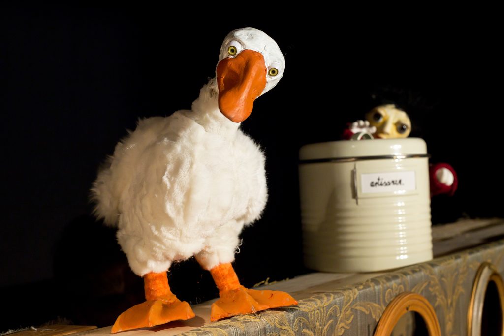 A white bird puppet with an orange beak and orange feet stands on a surface against a dark background. A light-colored bucket with a small skull resting on top is positioned to the right. The lighting highlights the texture of the feathers and the details of the objects.