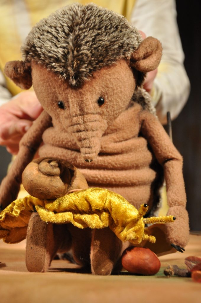 A hedgehog puppet with a brown body and a spiky back sits at a small table. A gold-colored fabric figure and a round brown object rest on the table. Hands operating the puppet are visible in the background.