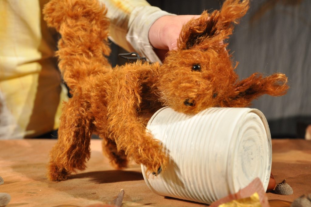 A brown animal puppet with shaggy fur leans against a white ribbed flowerpot. In the background, the hands of a person operating the puppet are visible. The scene is set on a tabletop with scattered natural materials and is lit with warm lighting.