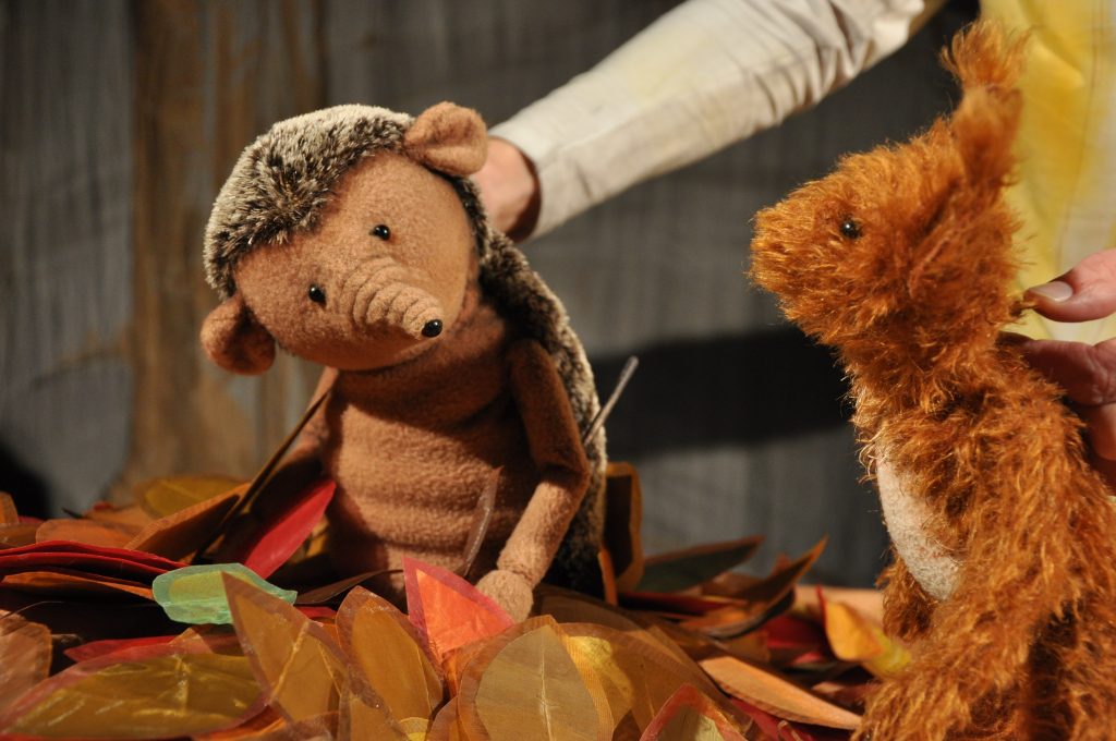 Eine Igelpuppe und eine braune Tierpuppe mit zotteligem Fell befinden sich auf einer Bühne zwischen dekorativen Blättern in Herbstfarben. Im Hintergrund sind die Hände der Puppenspielenden sichtbar. Die Szene ist warm beleuchtet und vor einer holzartigen Kulisse aufgebaut.
