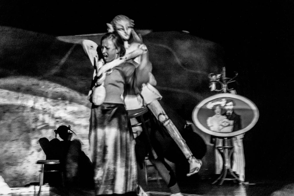 The blurry black-and-white picture shows a life-size puppet and a puppeteer. The puppeteer is carrying the puppet on her back and has a screaming expression on her face. In the background, there is a family photo in a round frame.