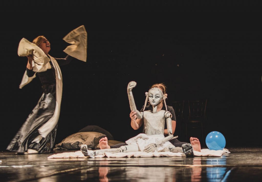 The picture shows two puppeteers and a puppet. The puppet is sitting on the floor with its arm raised. To the right of the puppet is a blue balloon. To the left of the puppet, the second puppeteer is dancing with golden fabrics.