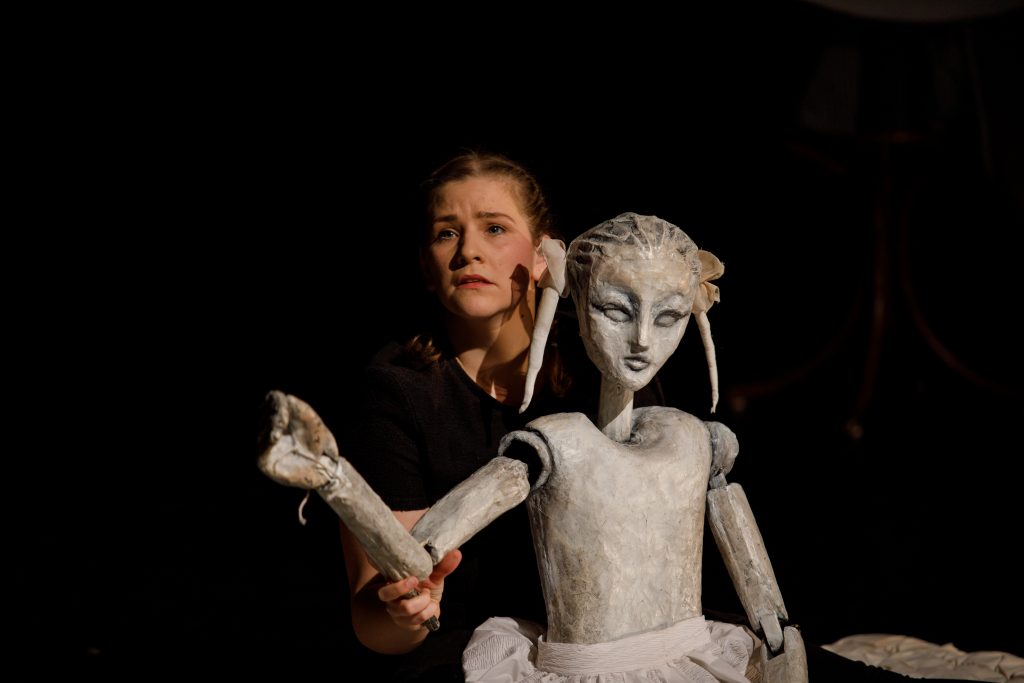 A close-up of a life-size puppet and a puppeteer. The puppet looks like a girl, and the puppeteer in the background has a sad expression on her face.