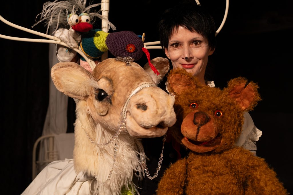 Several puppet figures are grouped against a dark background. In the foreground are a deer puppet with large antlers and a brown bear puppet. Behind them stands a person presenting the figures. A small colorful bird puppet is perched on the antlers. The scene is lit with warm lighting.